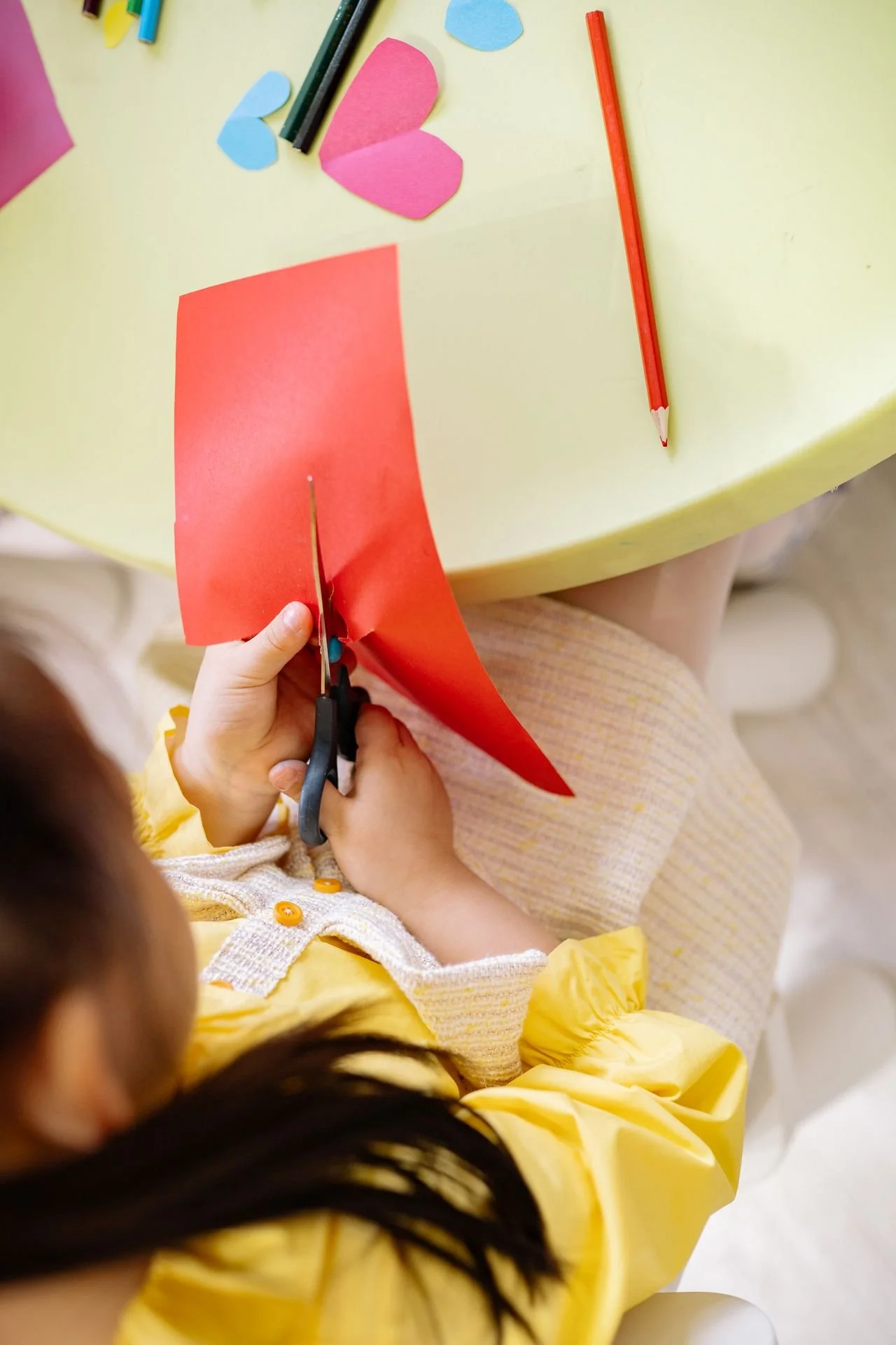 Ein Kind schneidet mit einer Schere rotes Papier, während auf einem gelben Tisch bmalen, Papierherzen und ein roder Bleistift liegen.