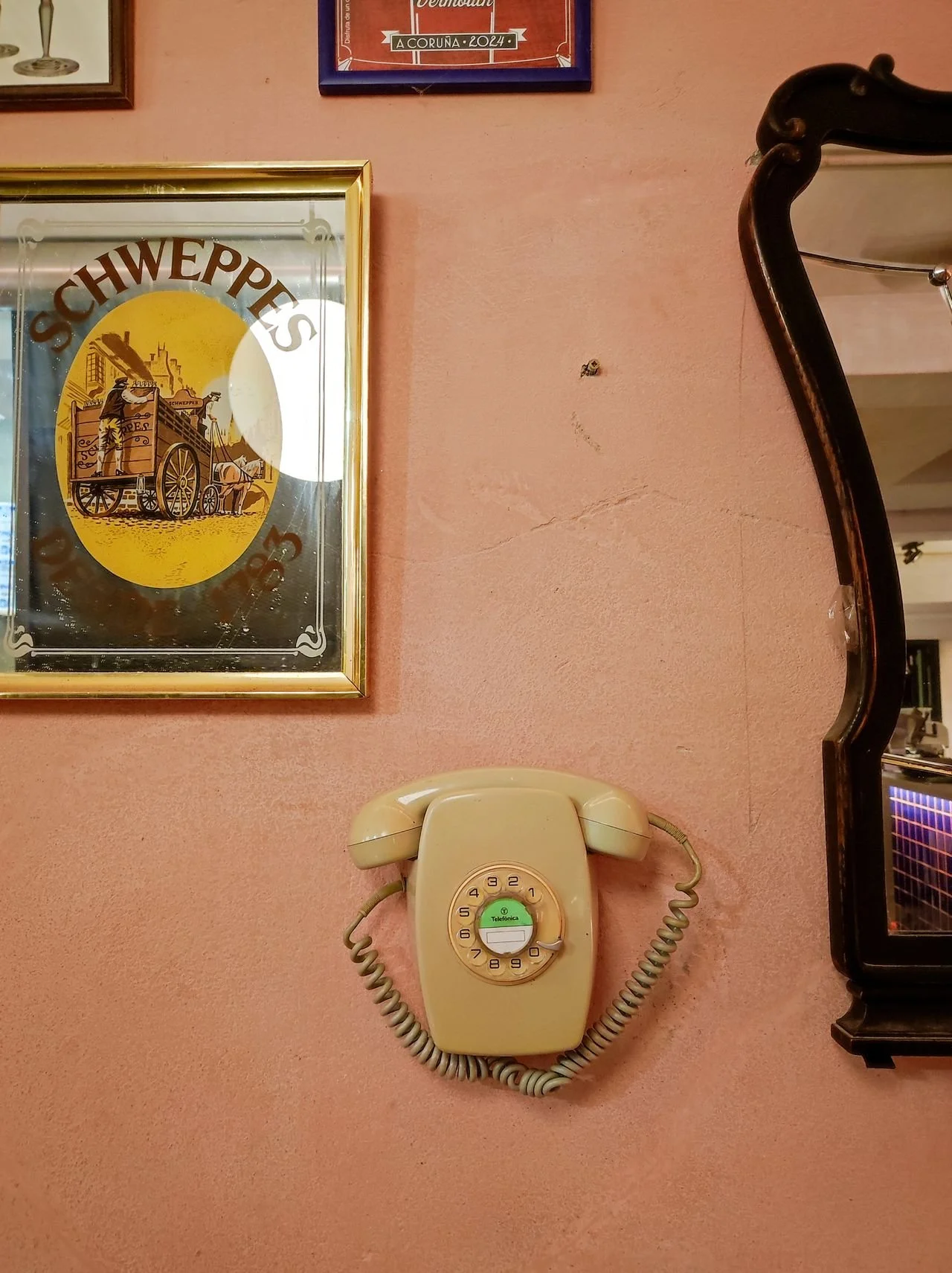 Ein alterer, hellgelber Wählautsprecher hängt an einer rosa Wand, darunter befindet sich ein hellgeltes Telefongerät. Auf der Wand hängen auch zwei gerahmte Bilder, eines mit einer Polizei- oder Marcha-Tafel und das andere mit einem Text über eine Veranstaltung im Jahr 2024 in A Coruña.