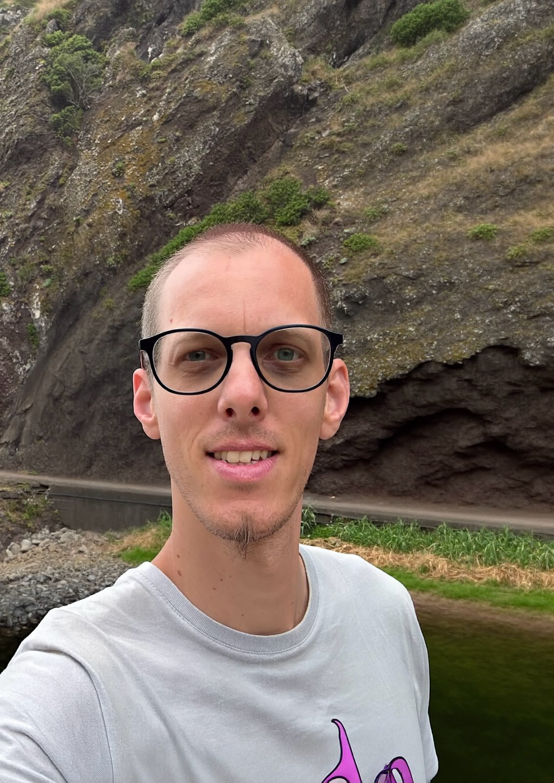 Ein Mann trägt eine schwarze Brille und ein weißes T-Shirt mit einer pinkfarbenen Grafik, steht vor einer felsigen und bewaldeten Landschaft.