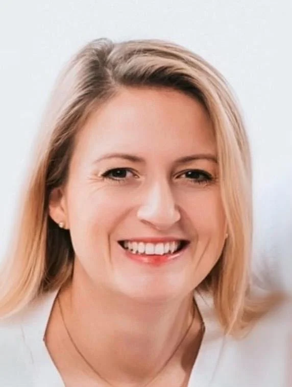 Ein Lächelndes Porträt einer Frau mit blondem Haar vor hellem Hintergrund.