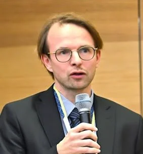 Ein Mann mit Brille, Anzug und Bluse hält ein Mikrofon und spricht bei einer Veranstaltung.