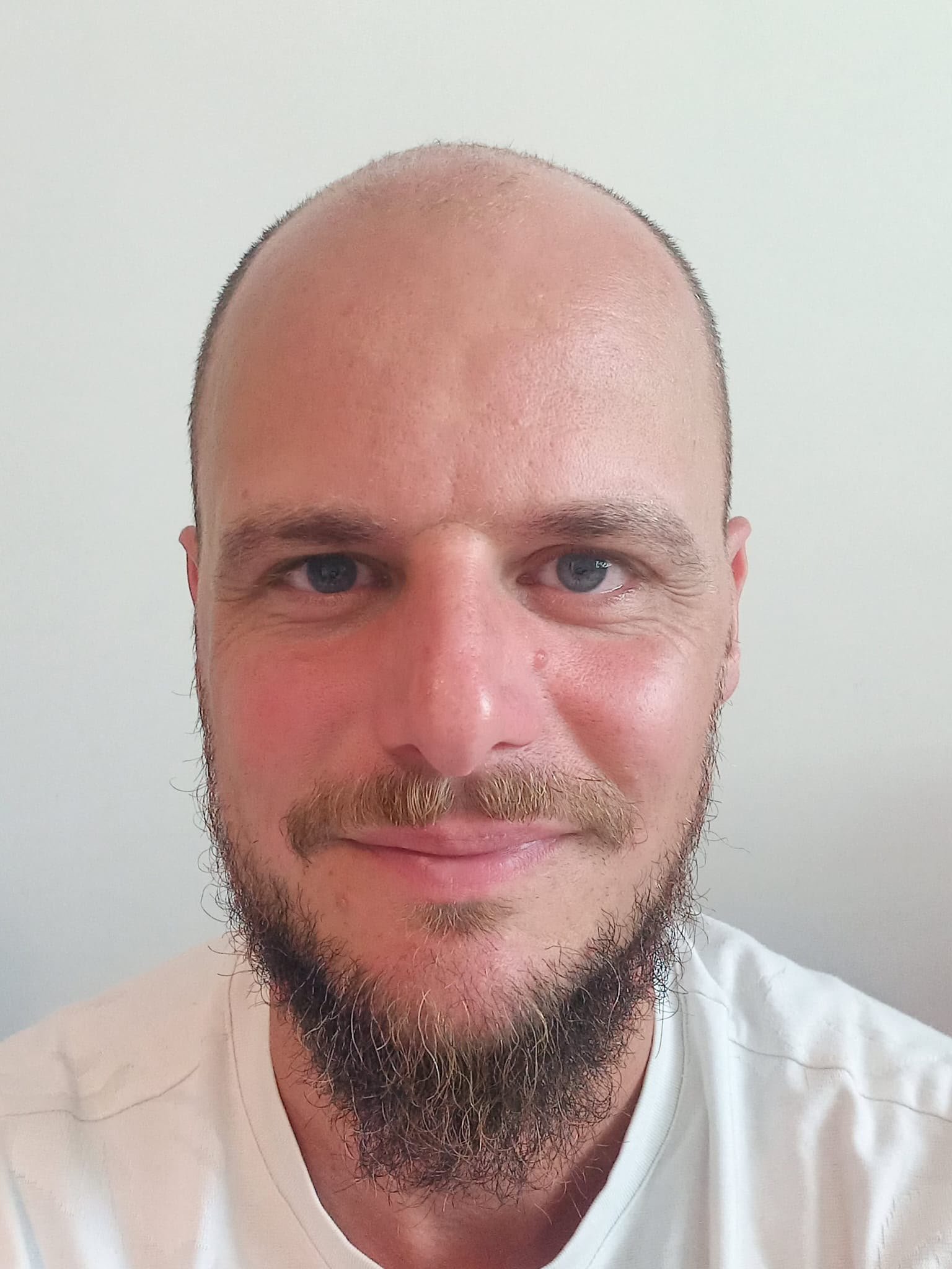 Ein Mann mit Glatze, blauen Augen, Bart und Vollmondgesicht, trägt ein weißes T-Shirt, steht vor einem hellen Wandhintergrund.