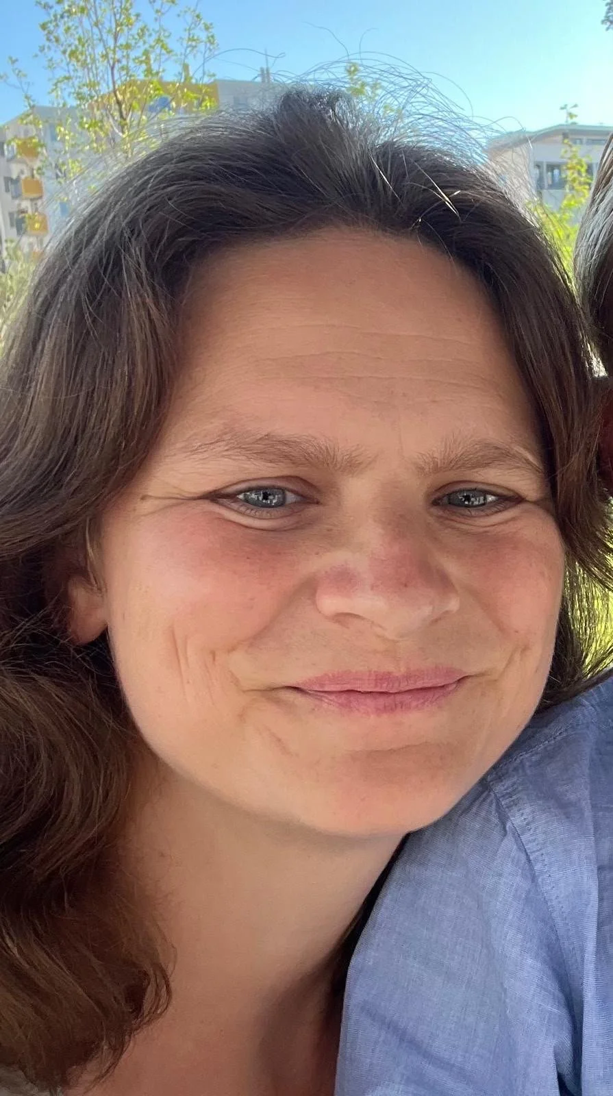 Nahaufnahme eines Lächelns einer Frau mit braunen, gewellten Haaren und blauen Augen, draußen bei Sonnenschein.