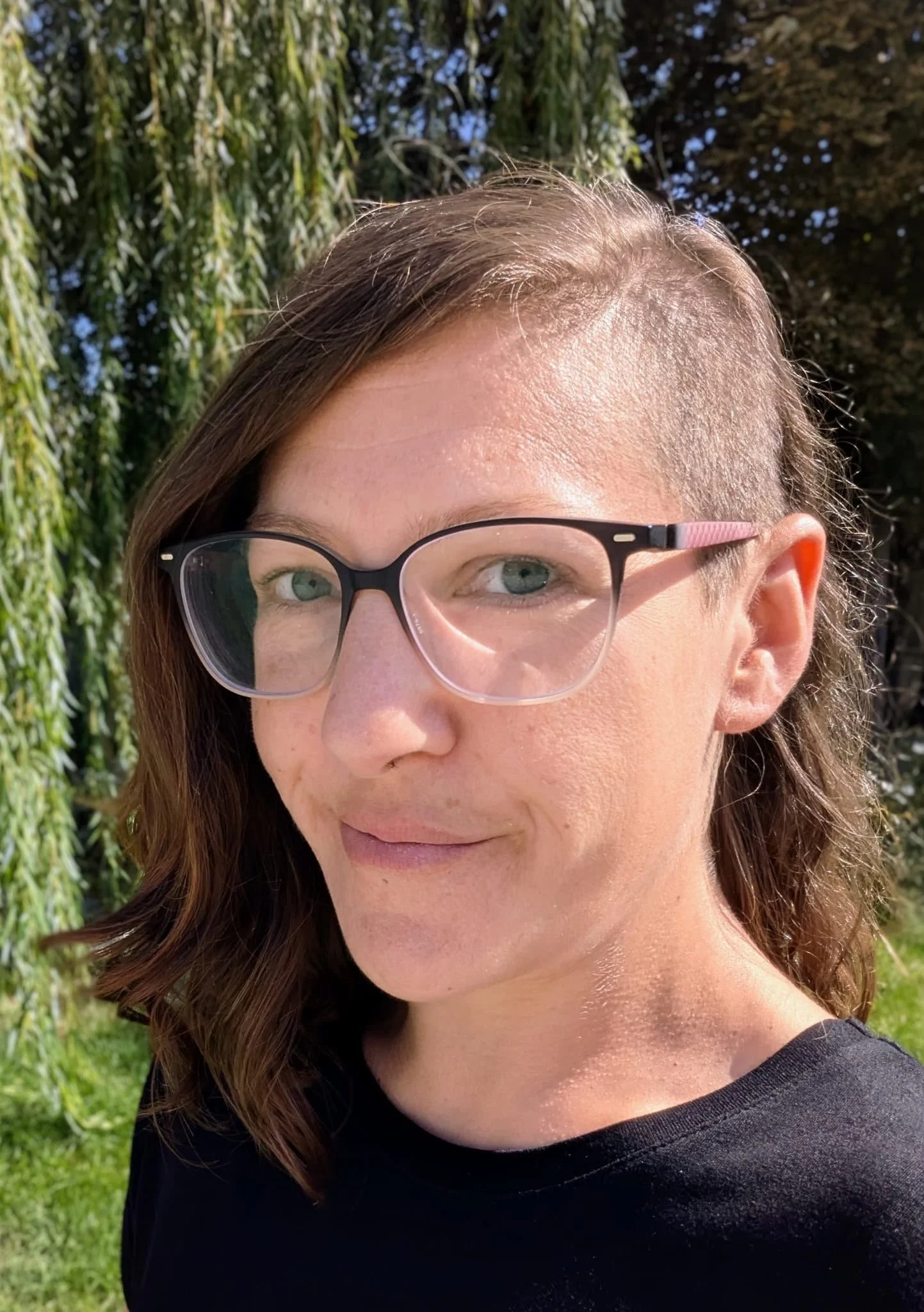 Nahaufnahme des Gesichts einer Frau mit Brille, braune Haare, die im Freien bei sonnigem Wetter steht, Hintergrund mit grünen Bäumen und Sonnenlicht.