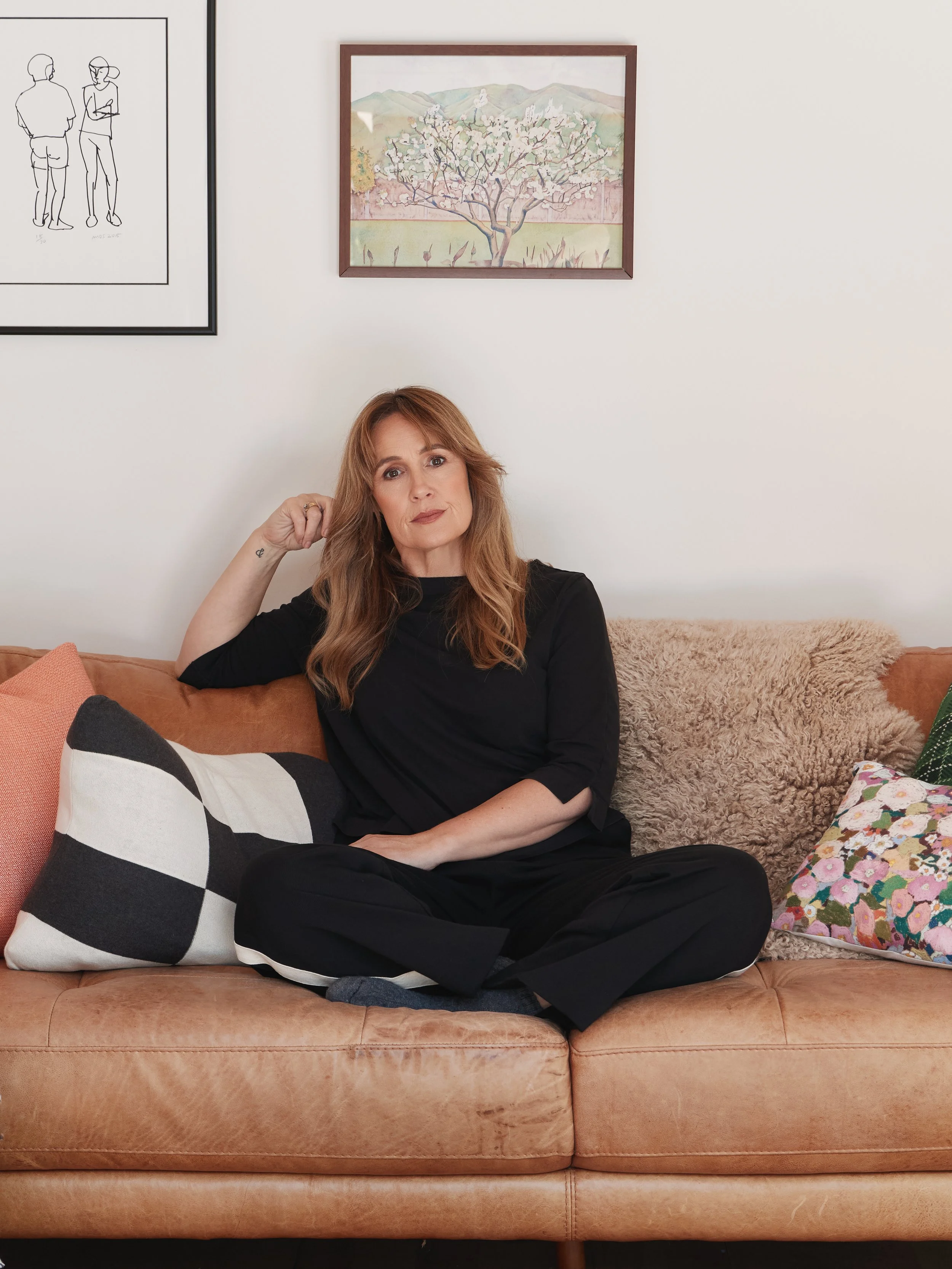 Woman in black clothing sitting cross-legged on a brown leather sofa beneath framed artwork.