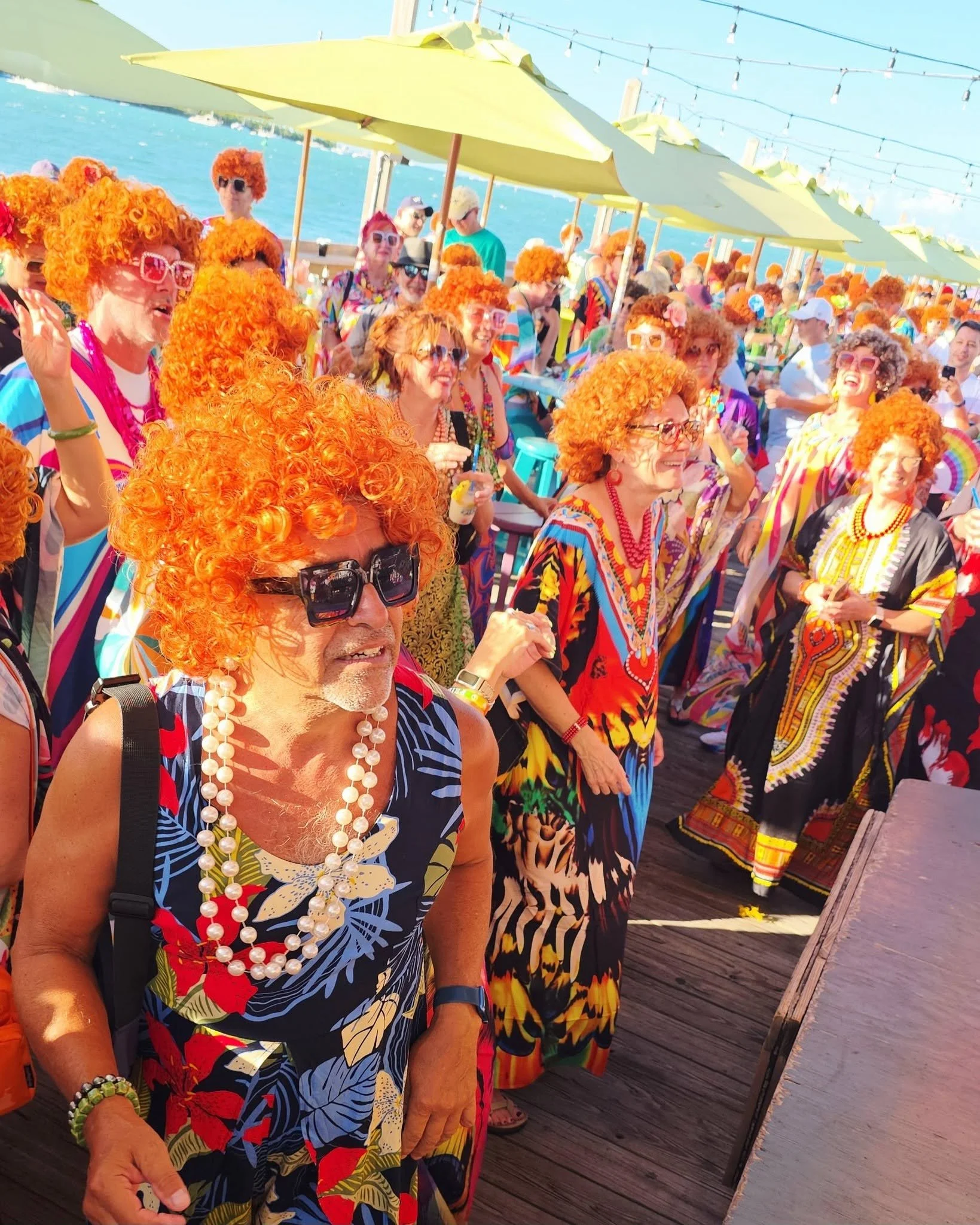 Dozens of people wearing colorful caftans wearing orange wigs on a pier with umbrellas.