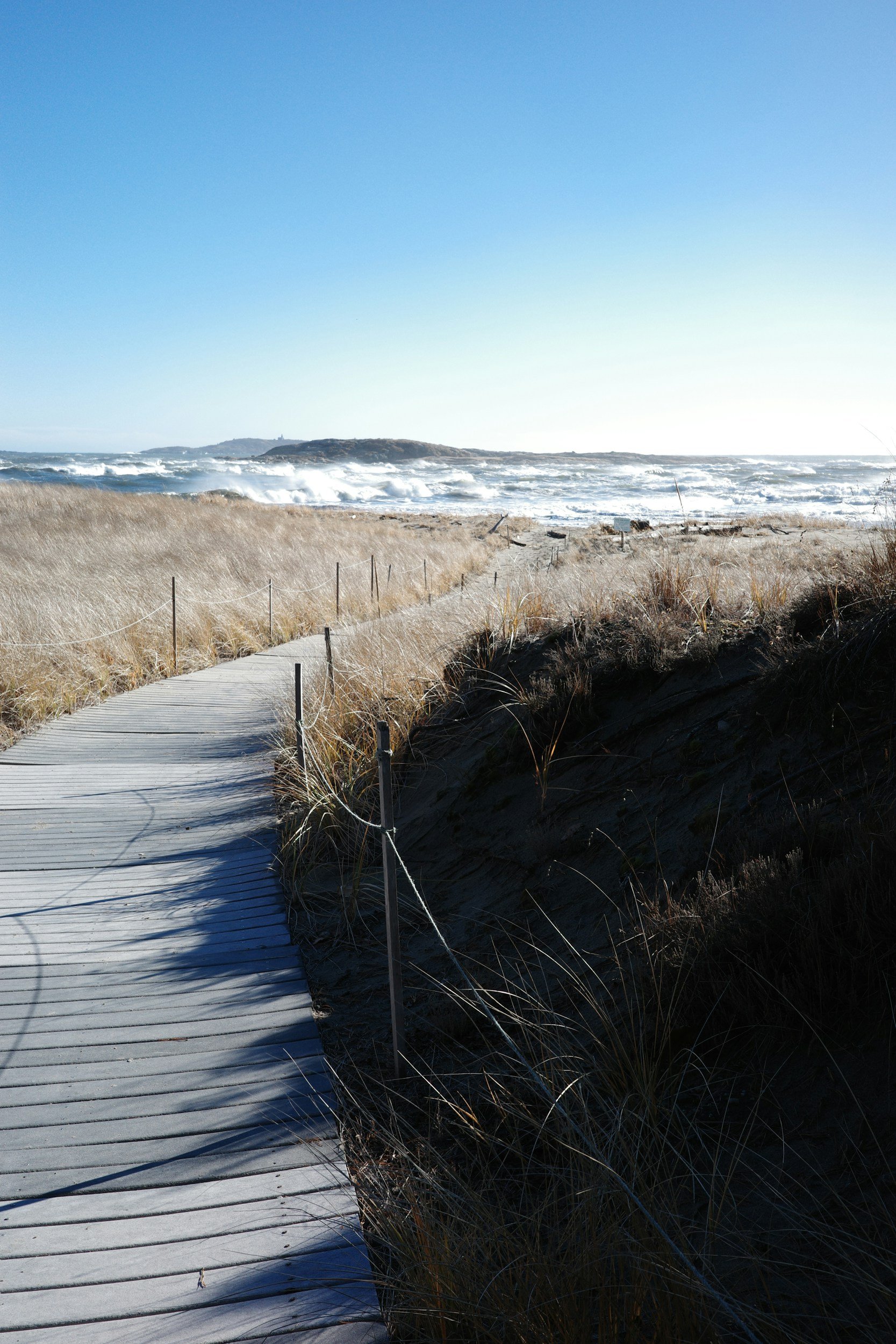 Wooden boardwalk through coastal dunes leading to a rocky island and crashing ocean waves