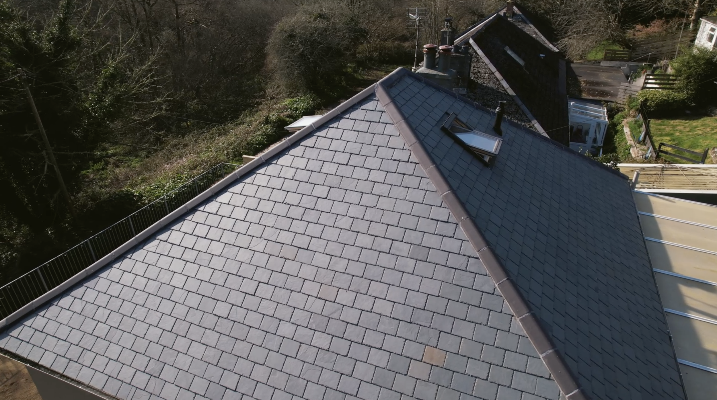 An aerial view of a house roof covered with gray shingles, with a skylight window and several chimneys, surrounded by trees and neighboring houses.
