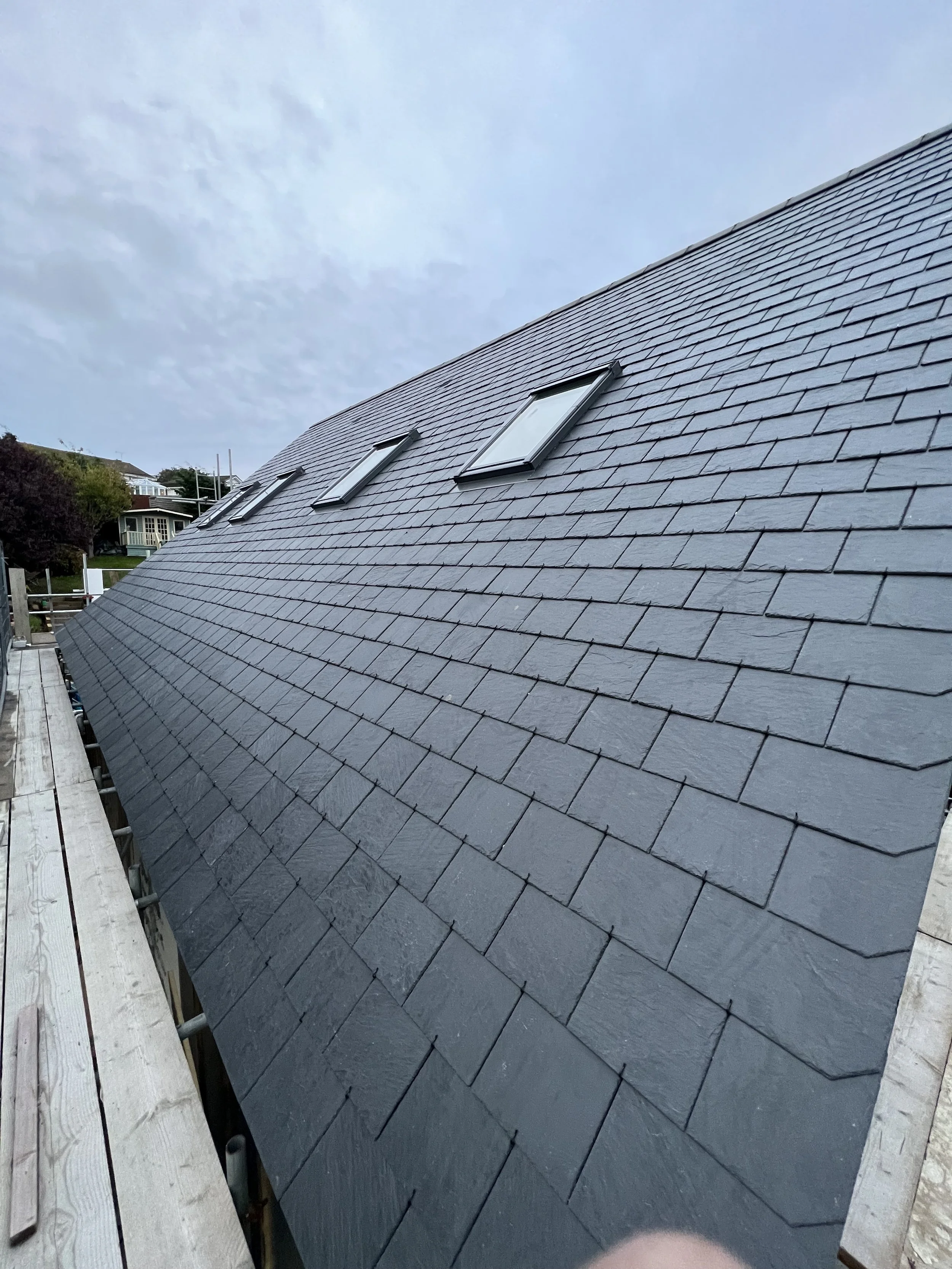 Sloped roof with slate shingles and three skylights, with a background of a blue sky and distant houses.
