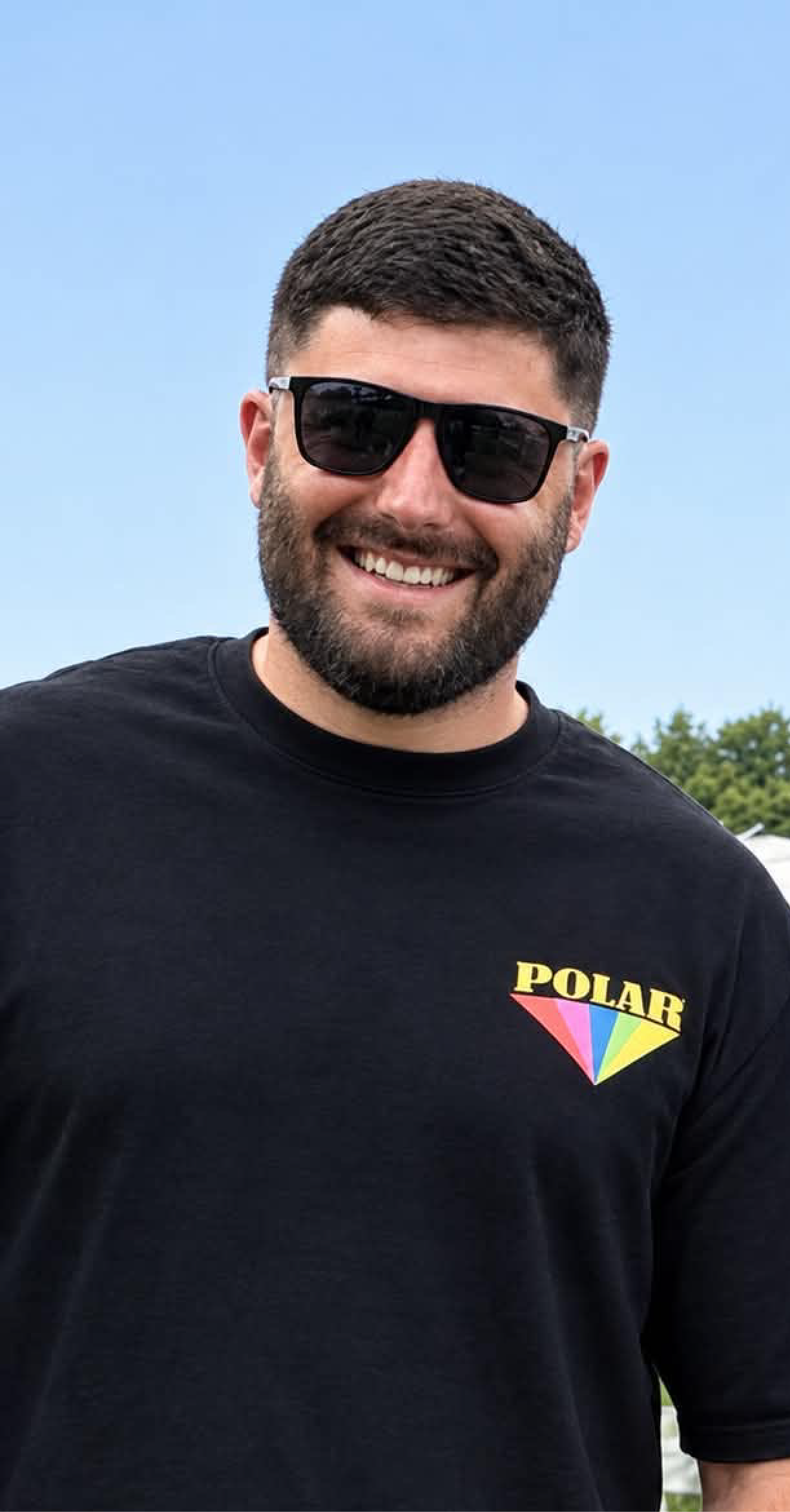 A man with short dark hair and a beard wearing sunglasses and a black t-shirt with a rainbow-colored logo reading 'POLAR'.