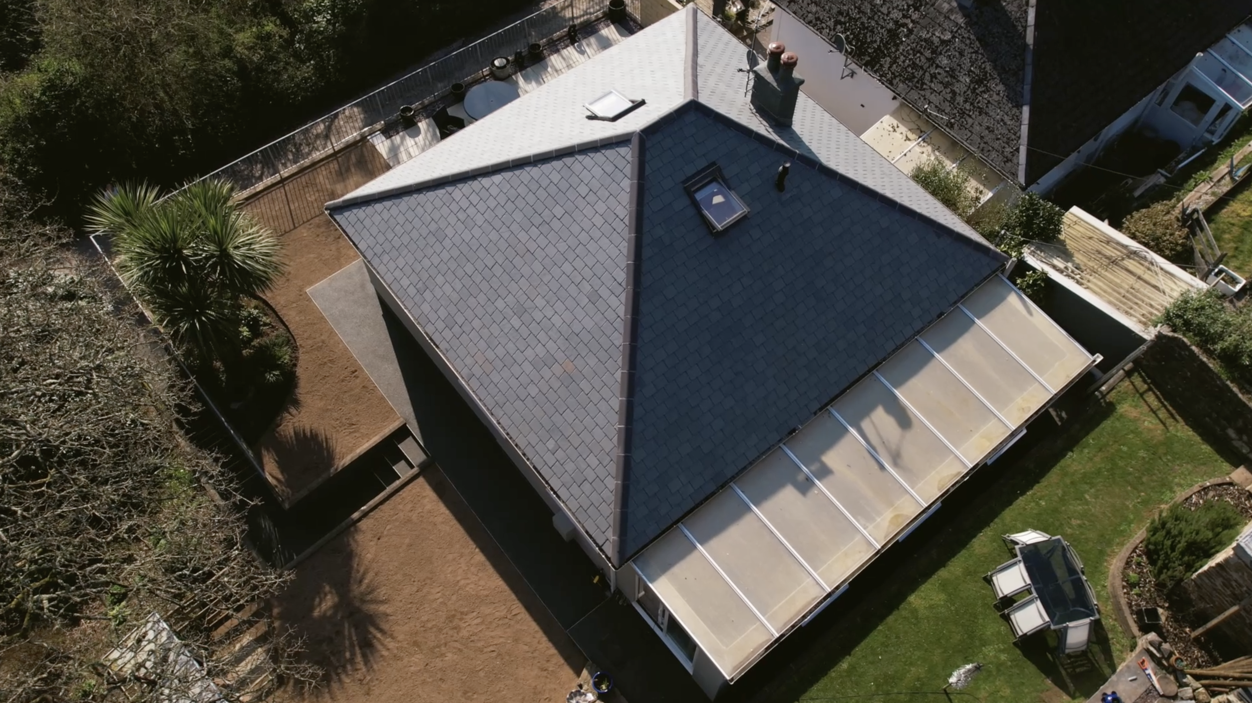An aerial view of a house with a gray shingled roof, surrounded by trees and a fenced backyard.