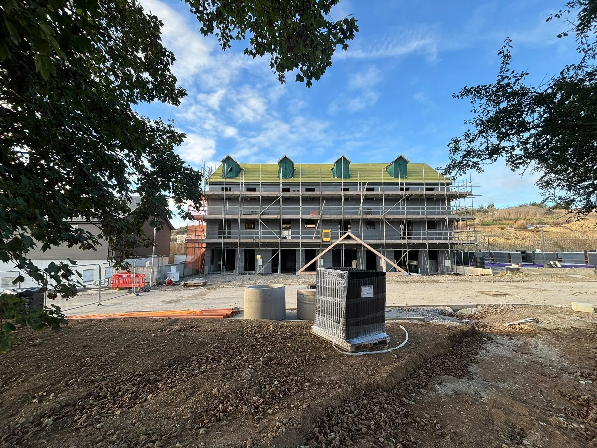 Construction site with a new building under construction, scaffolding around the structure, and trees framing the scene with a blue sky and clouds overhead.