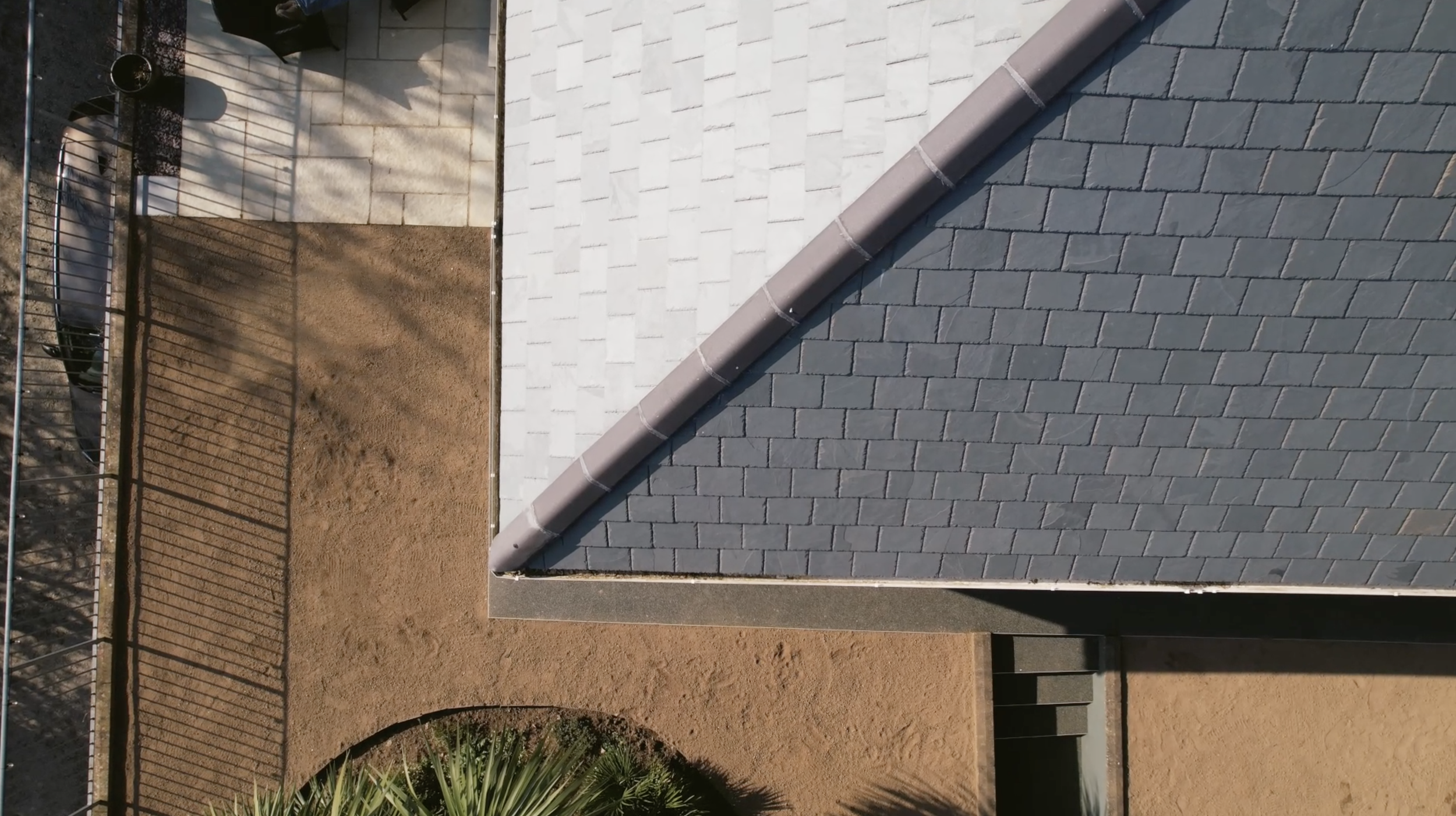 An aerial view of a house roof with gray shingles, a metal gutter running along the edge, and a small yard enclosed by a black metal fence. The yard has a dirt surface, and there are shadows cast by nearby trees and structures.