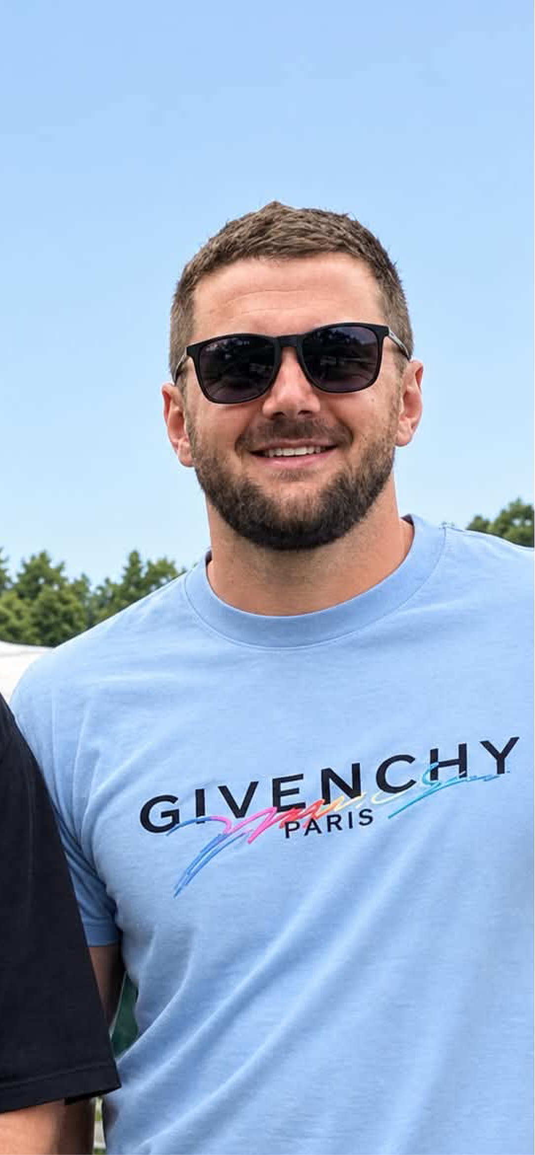 A smiling man wearing black sunglasses and a light blue Givenchy Paris T-shirt outdoors during daytime.