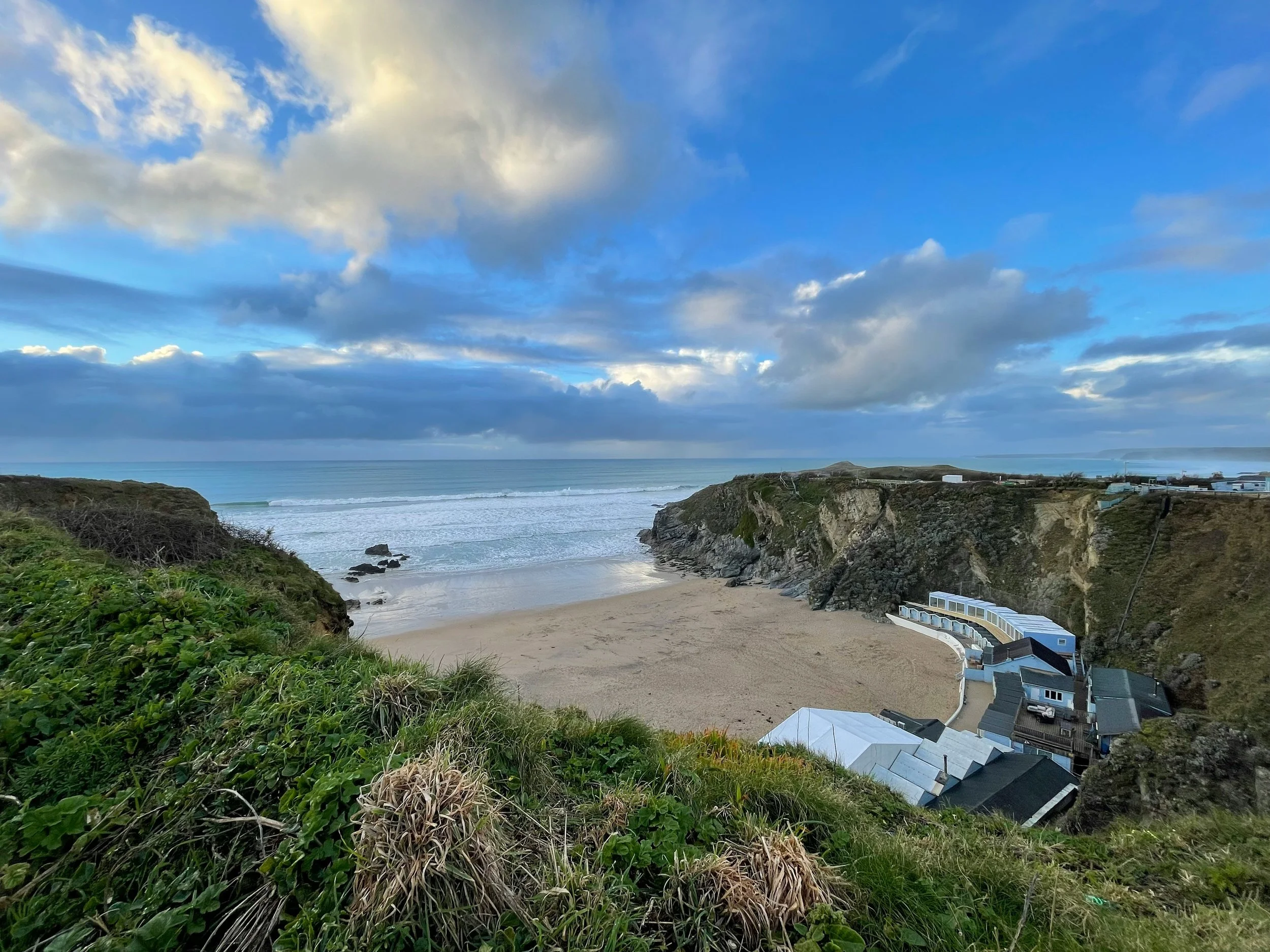View of a coastal landscape with a sandy beach, rocky cliffs, the ocean, and houses along the shoreline, under a partly cloudy sky.