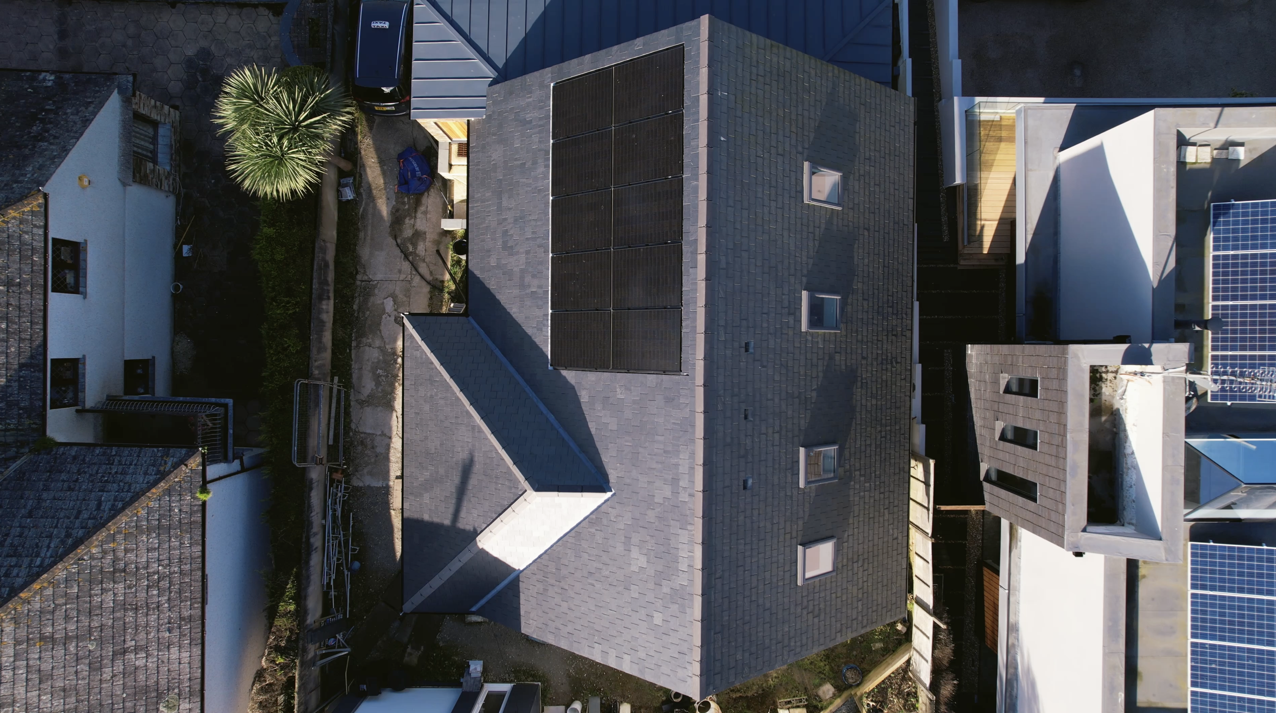 An aerial view of rooftops of several houses, some with solar panels, surrounded by trees and neighboring buildings.