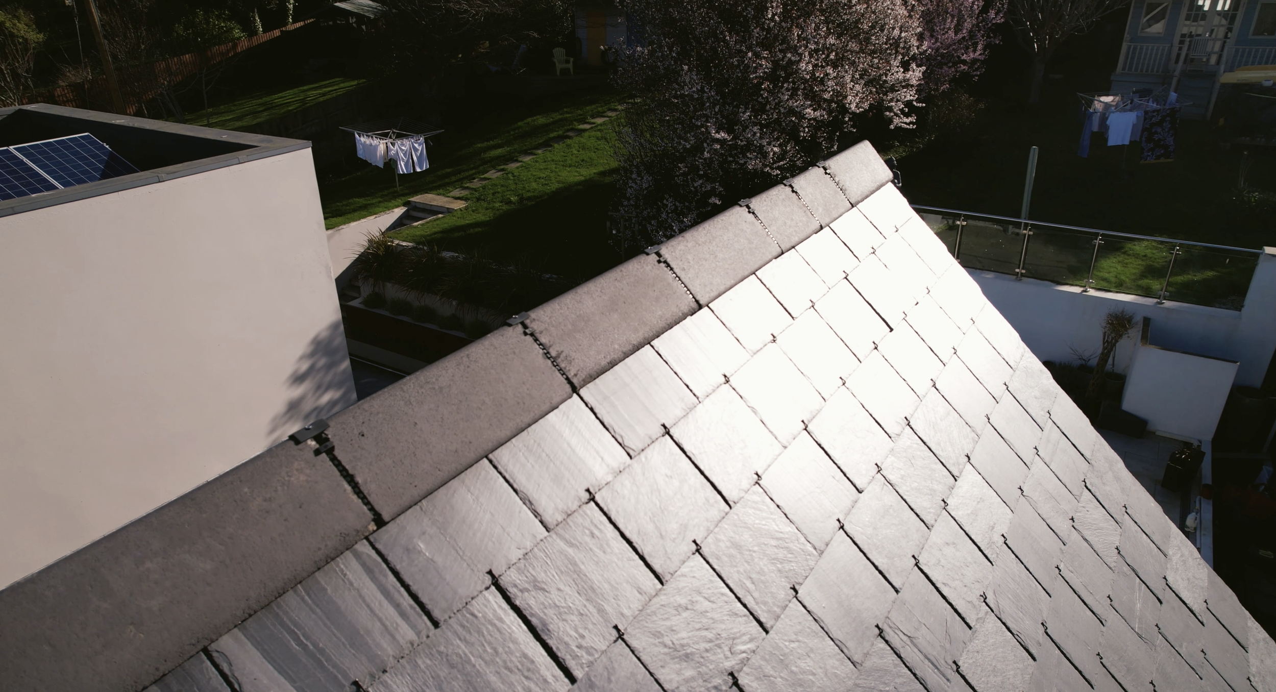 A house roof with solar panels in a backyard, a tree with pink blossoms, laundry hanging on a clothesline, a white fence, and a small seating area with a white chair.