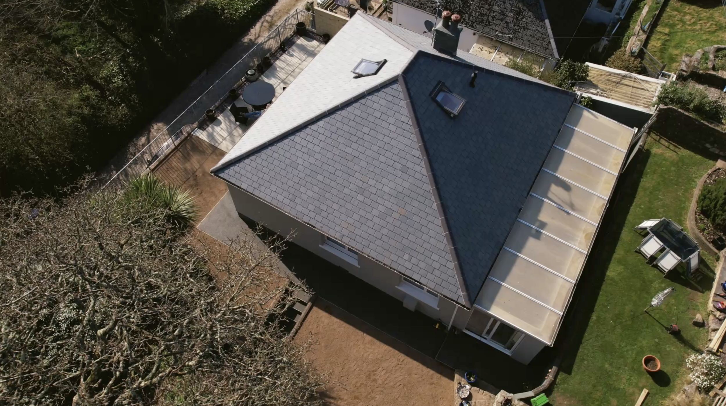 An aerial view of a house with a dark gray roof and a small backyard patio with seating, surrounded by trees and grass.