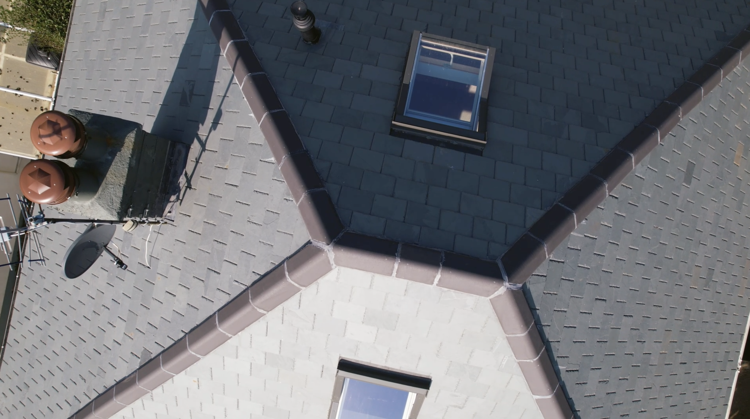 Aerial view of a house roof showing a skylight window, two brown ventilation pipes, an air conditioning unit, and a satellite dish.