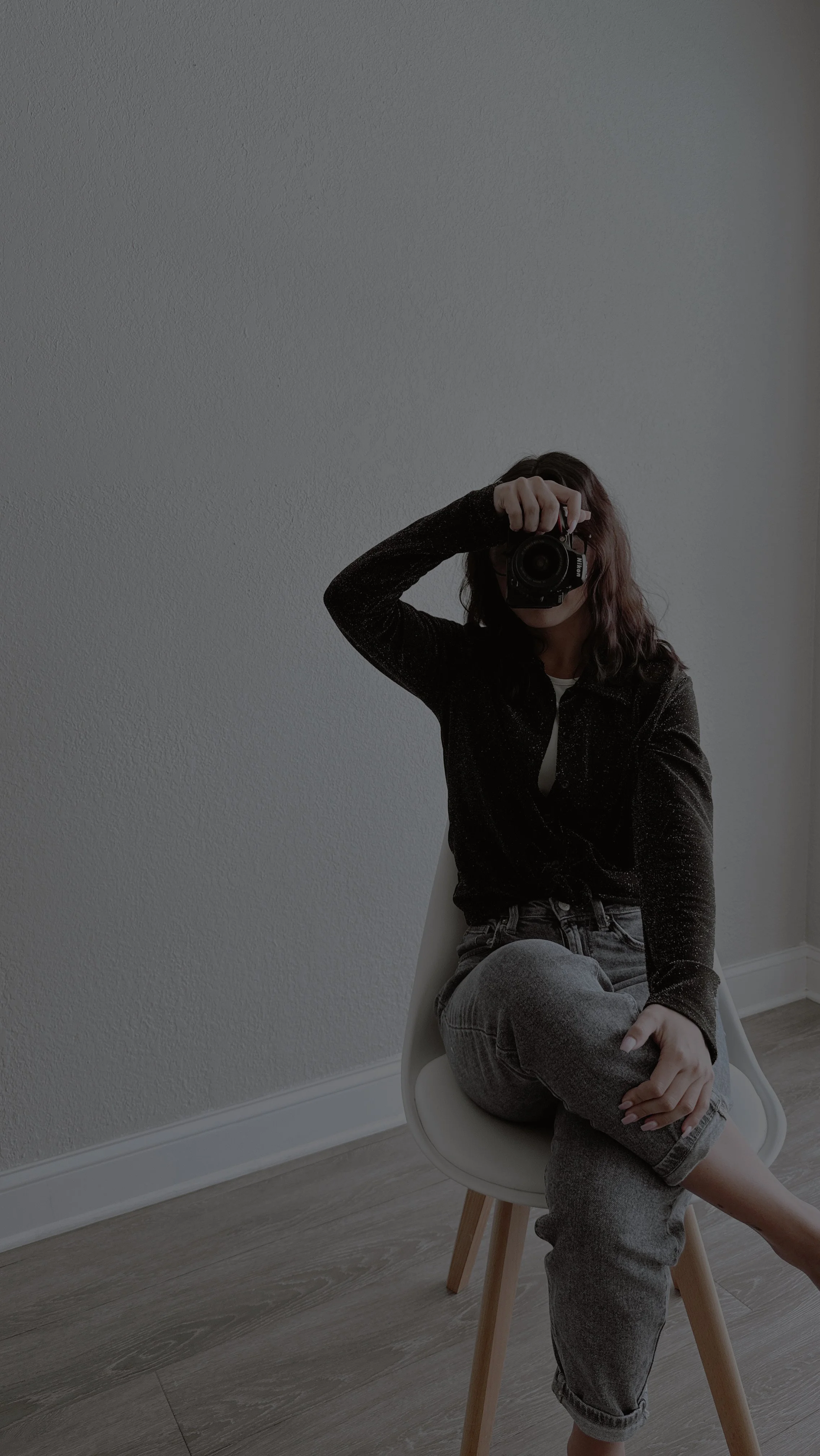 Person in a dark sweater and gray jeans sitting in a chair and photographing themselves with a Nikon camera.