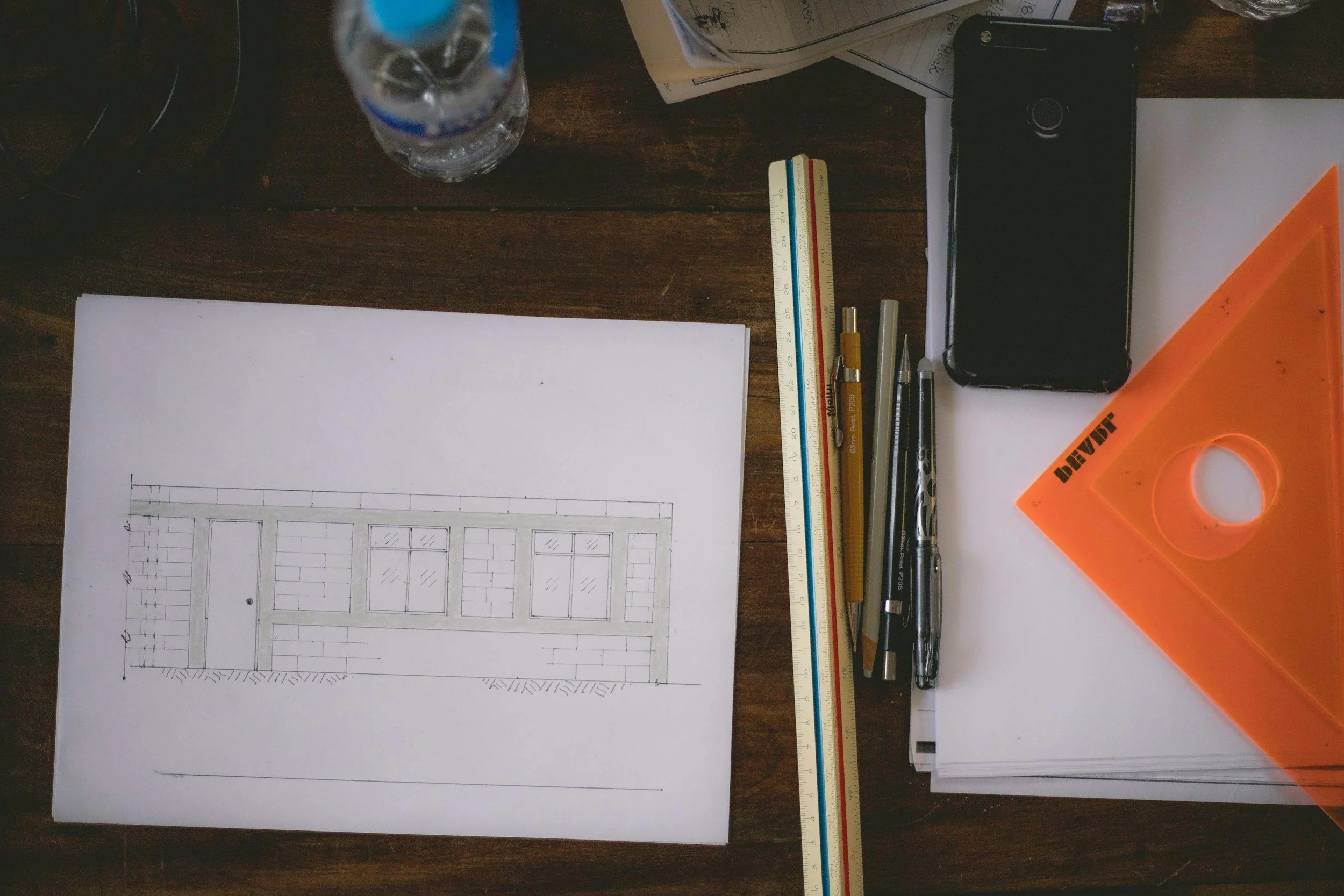 Mesa de dibujo con plano arquitectónico de una fachada, regla, lápices, bolígrafos, escuadra naranja, cuaderno y botella de agua.