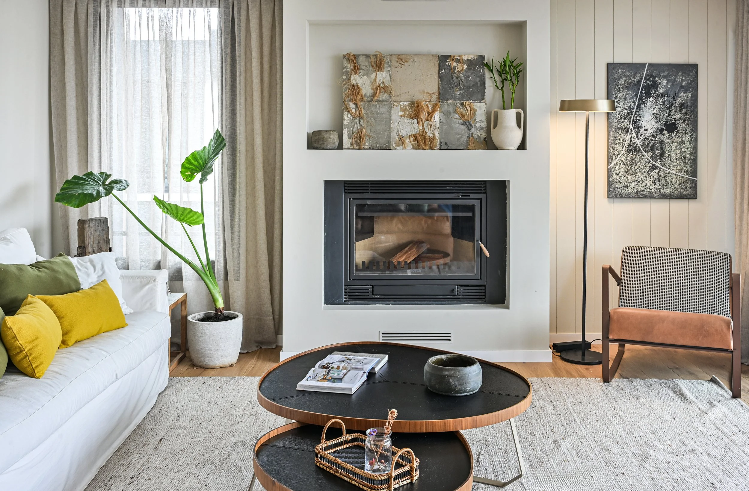 Sala de estar moderna y luminosa con chimenea, sofá blanco, planta tropical, mesas de centro, lámpara de pie y sillón. Esto pertenece a la expocisión en Casadeco Angra Open House 2026.