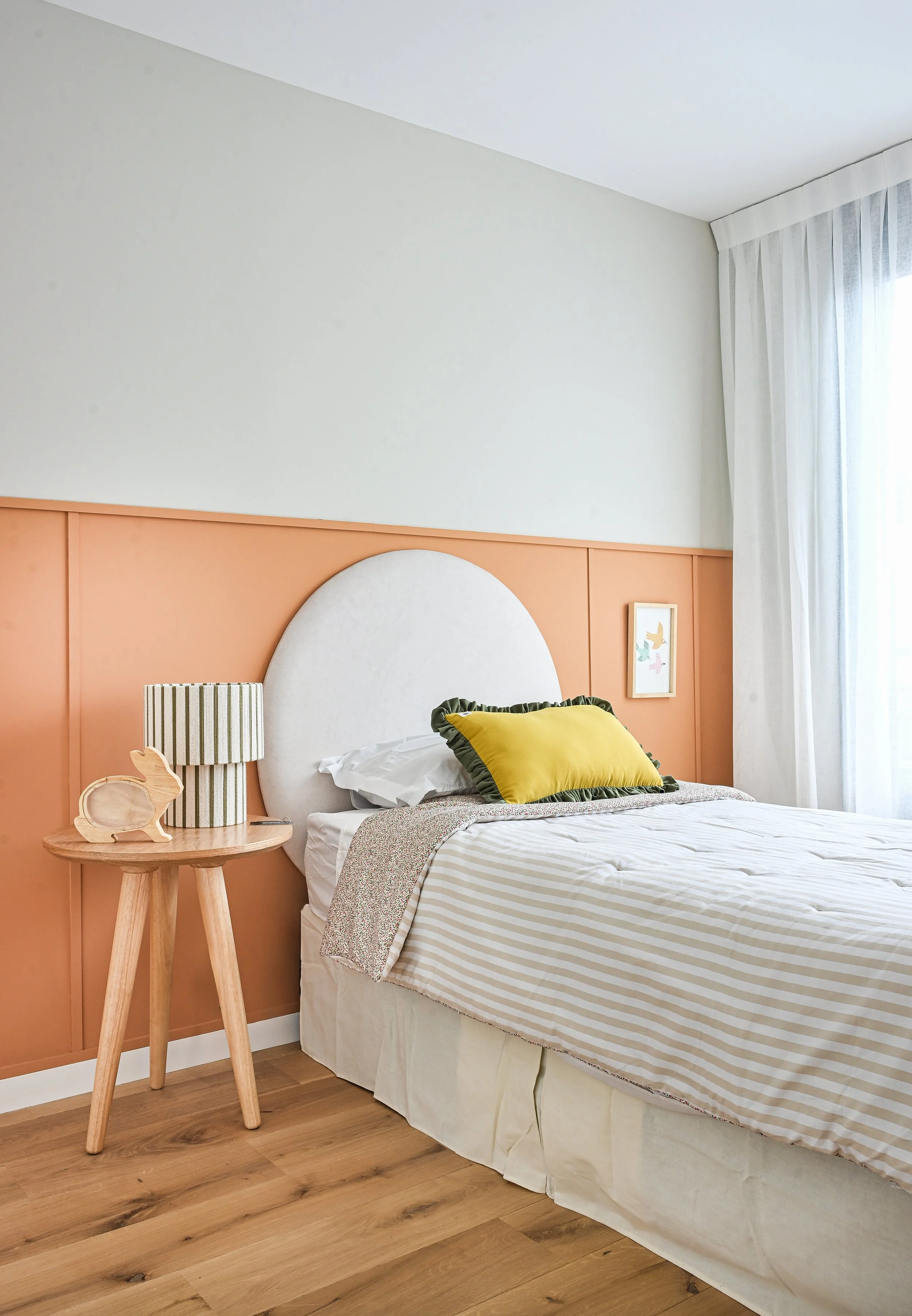 Dormitorio infantil moderno con cama, cabecera semicircular, almohada amarilla, mesa auxiliar de madera y cortinas blancas.