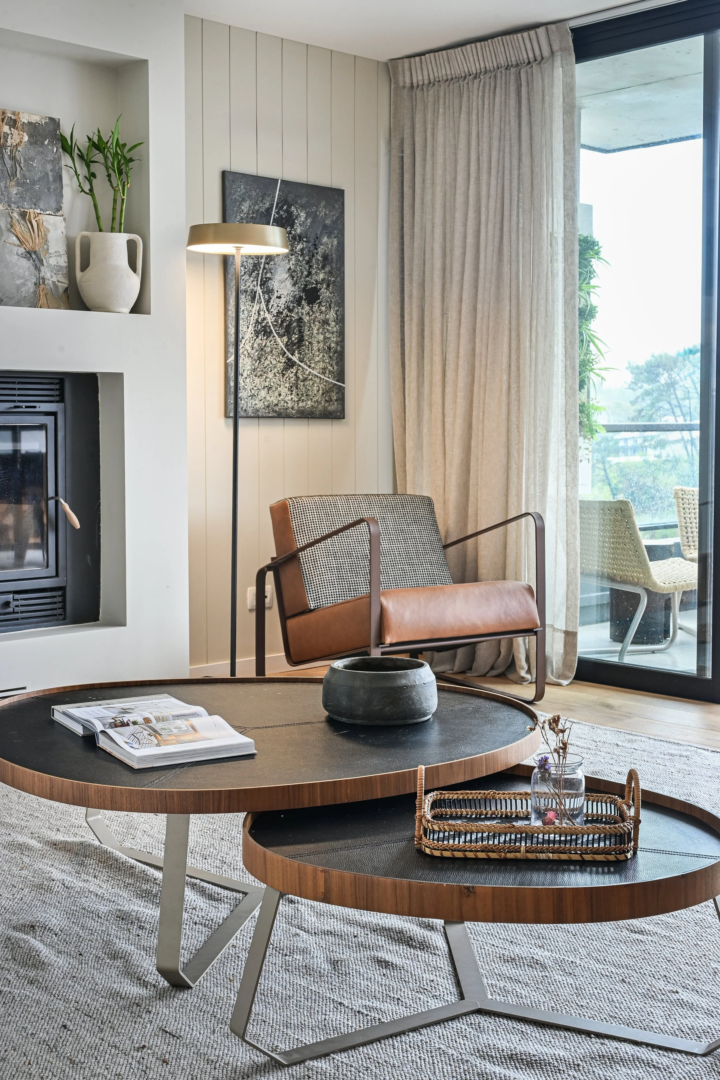 Sala moderna y luminosa con chimenea, sillón de cuero, mesas de centro redondas, planta de bambú, cuadros decorativos y ventanales con cortinas. Esto pertenece a la expocisión en Casadeco Angra Open House 2026.
