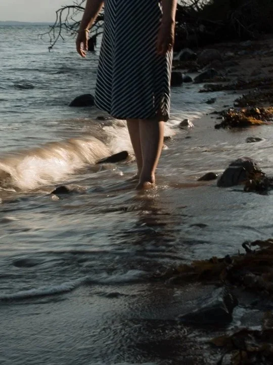 En person står i vandkanten på en strand, iført en sort og hvid stribet kjole, med tæer i vandet.
