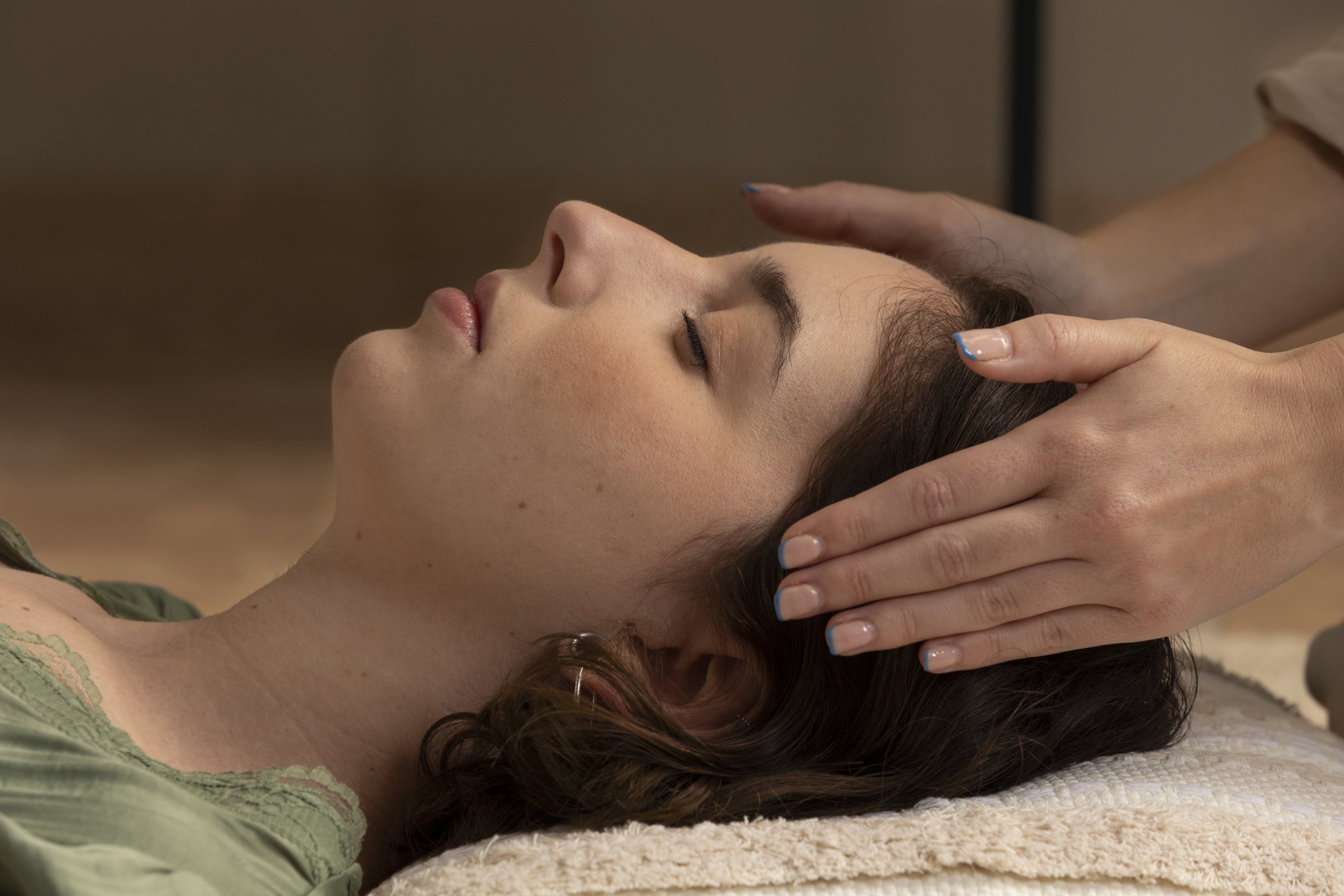En kvinde får en afslappende massage i hovedet og nakken, liggende på en blød, beige måtte.