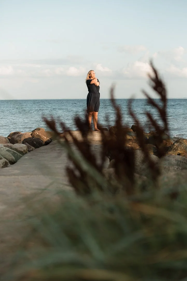Kvinde står på en stenbro ud mod havet med hænderne foldede, omgivet af sten og uklar vegetation i fronten.