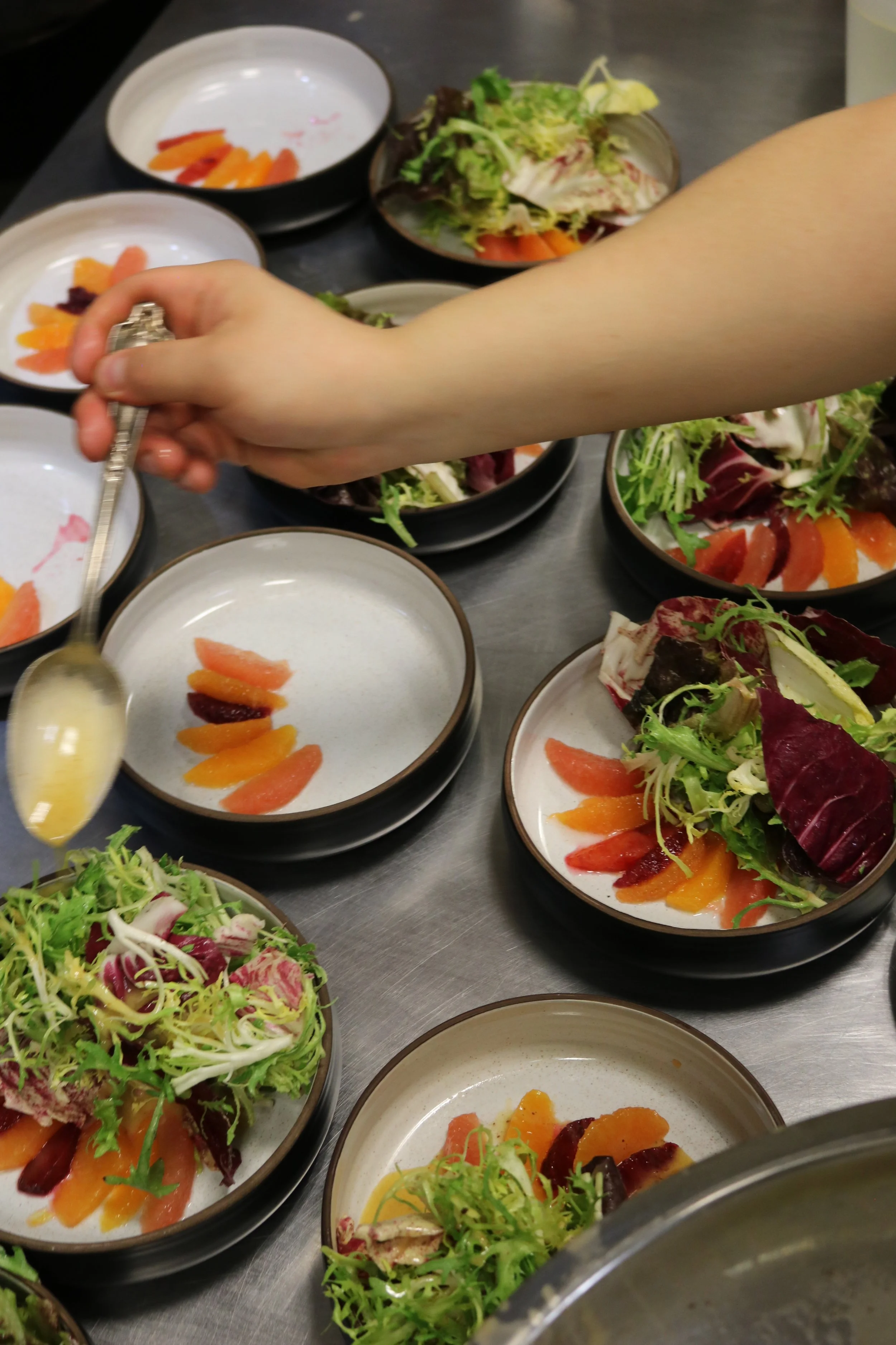 A person's arm and hand holding a spoon with salad bowls and plates of mixed greens, lettuce, and tomato slices on a stainless steel surface.