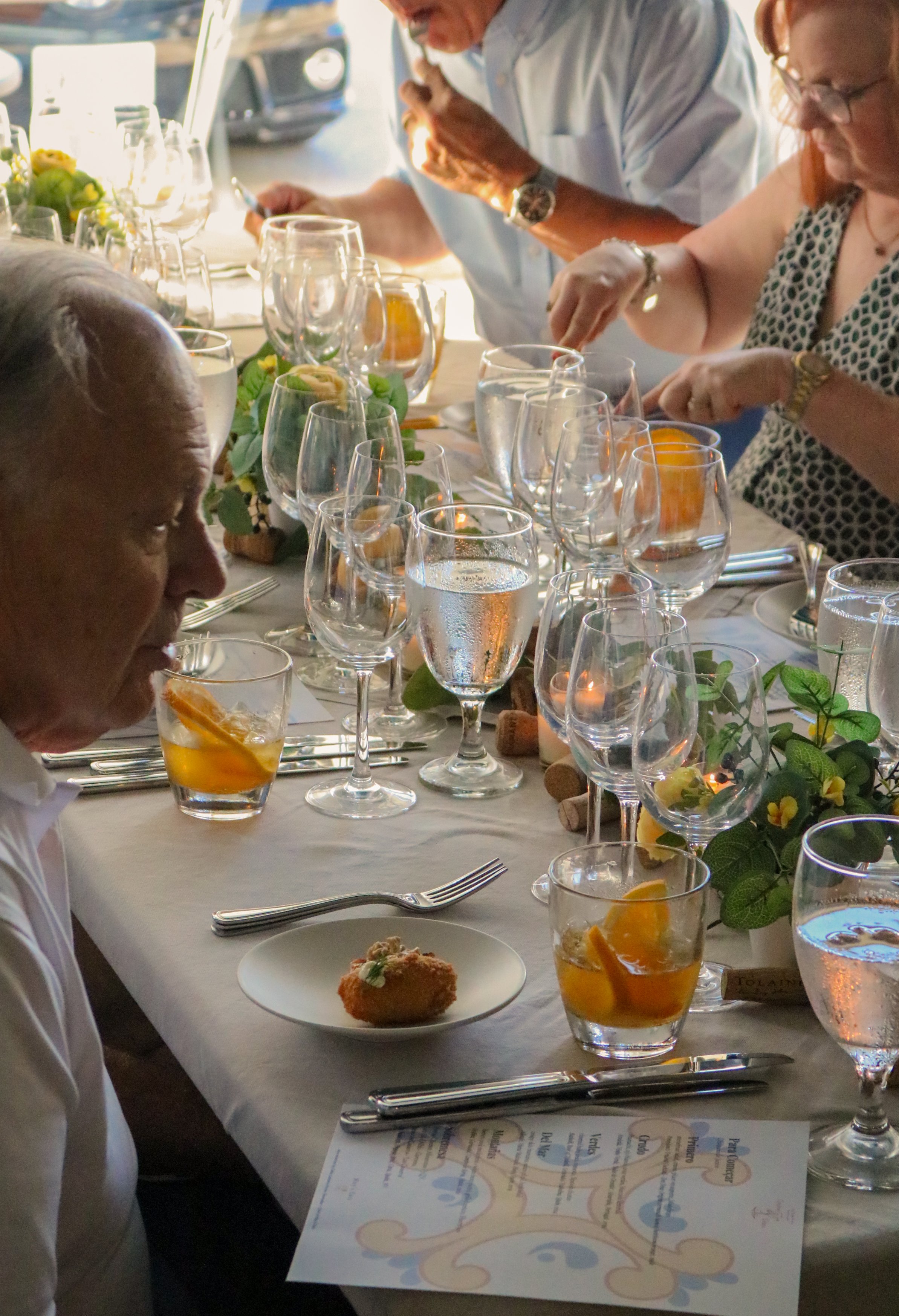 Guests seated at a long dining table with white tablecloth, adorned with glasses of water, orange drinks, decorative greenery, and small plates of appetizers. The people are engaged in conversation and appear to be enjoying a meal, possibly at a celebration or special event.