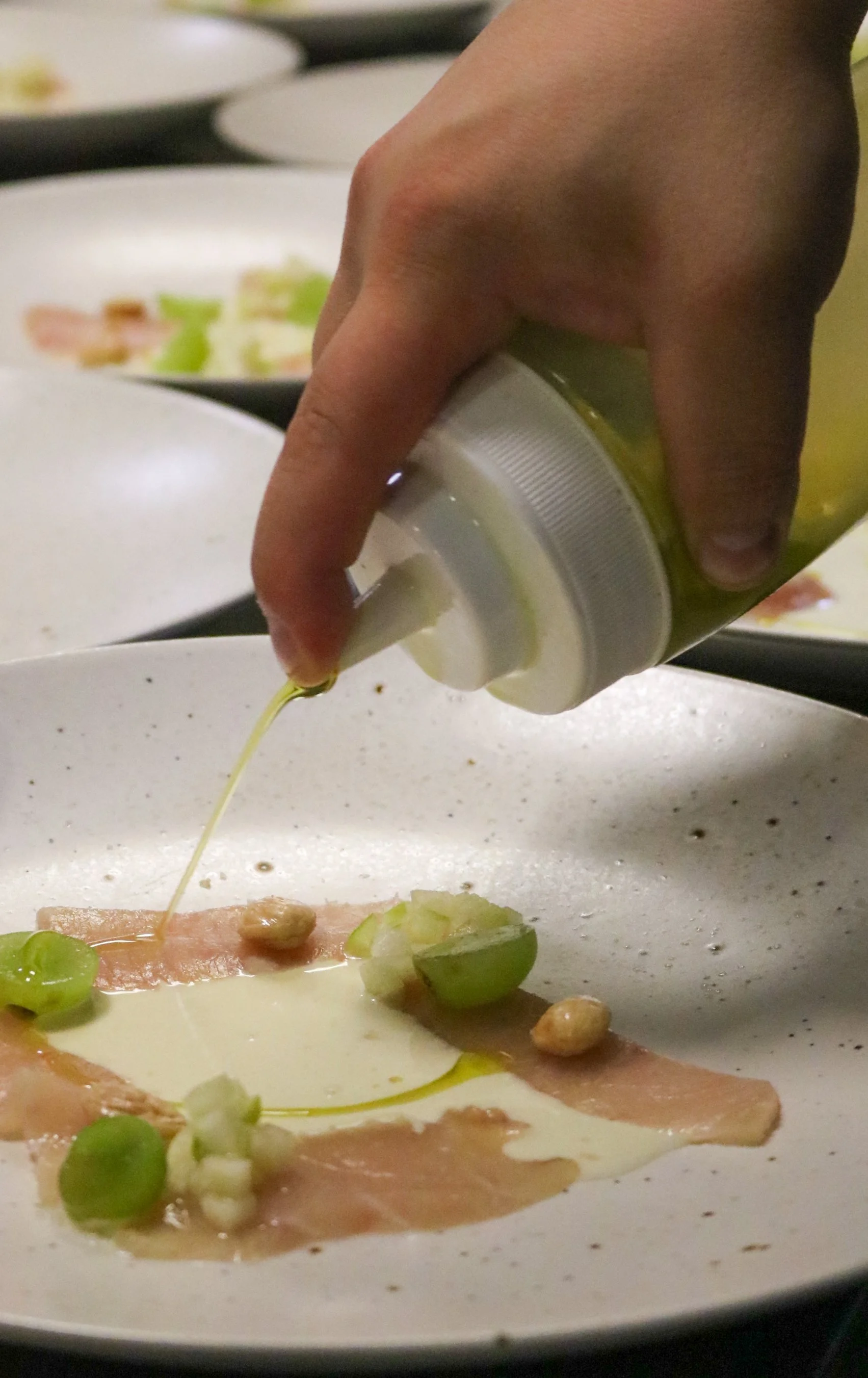 Person pouring olive oil onto a dish that includes sliced green grapes, thin slices of meat, and nuts in a white bowl.