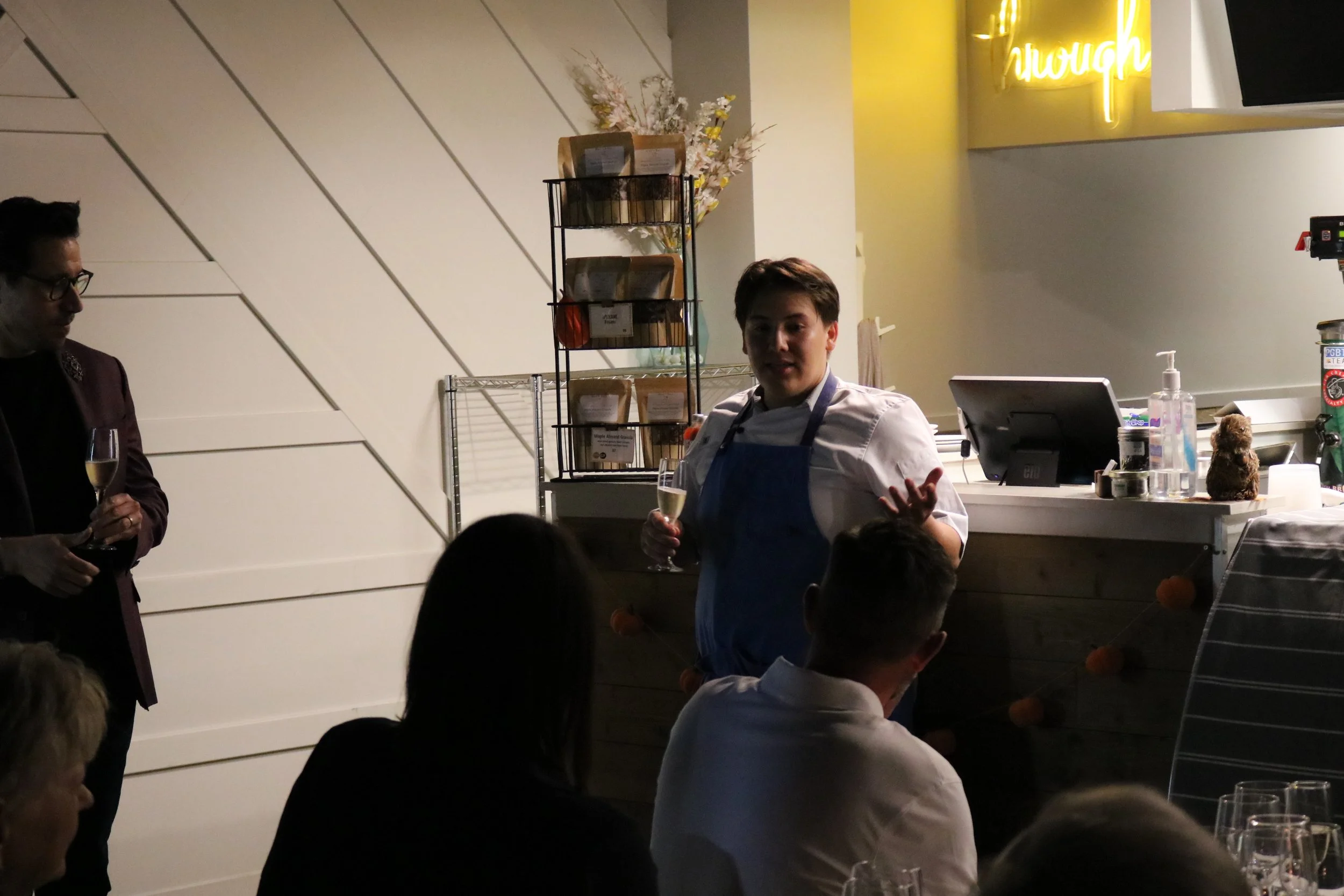 A woman in a white shirt and blue apron is speaking to a group in a restaurant or event space, holding a glass of champagne. To her left, a man in glasses and a dark jacket holds a similar glass. The background features a wall with floral decorations