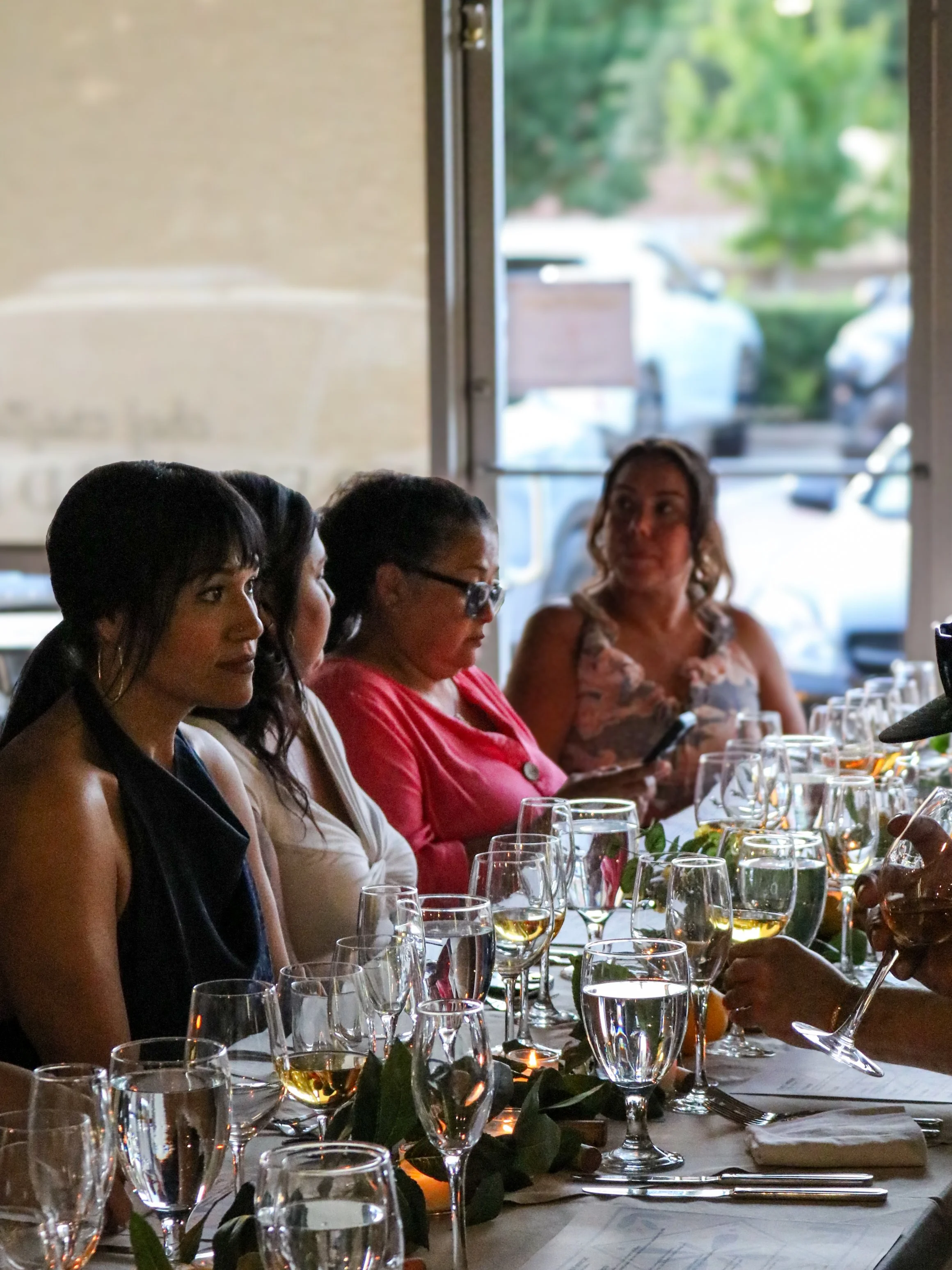 People sitting at a formal dining table with wine glasses and table settings, engaged in conversation in a restaurant with a large window showing outdoor greenery.