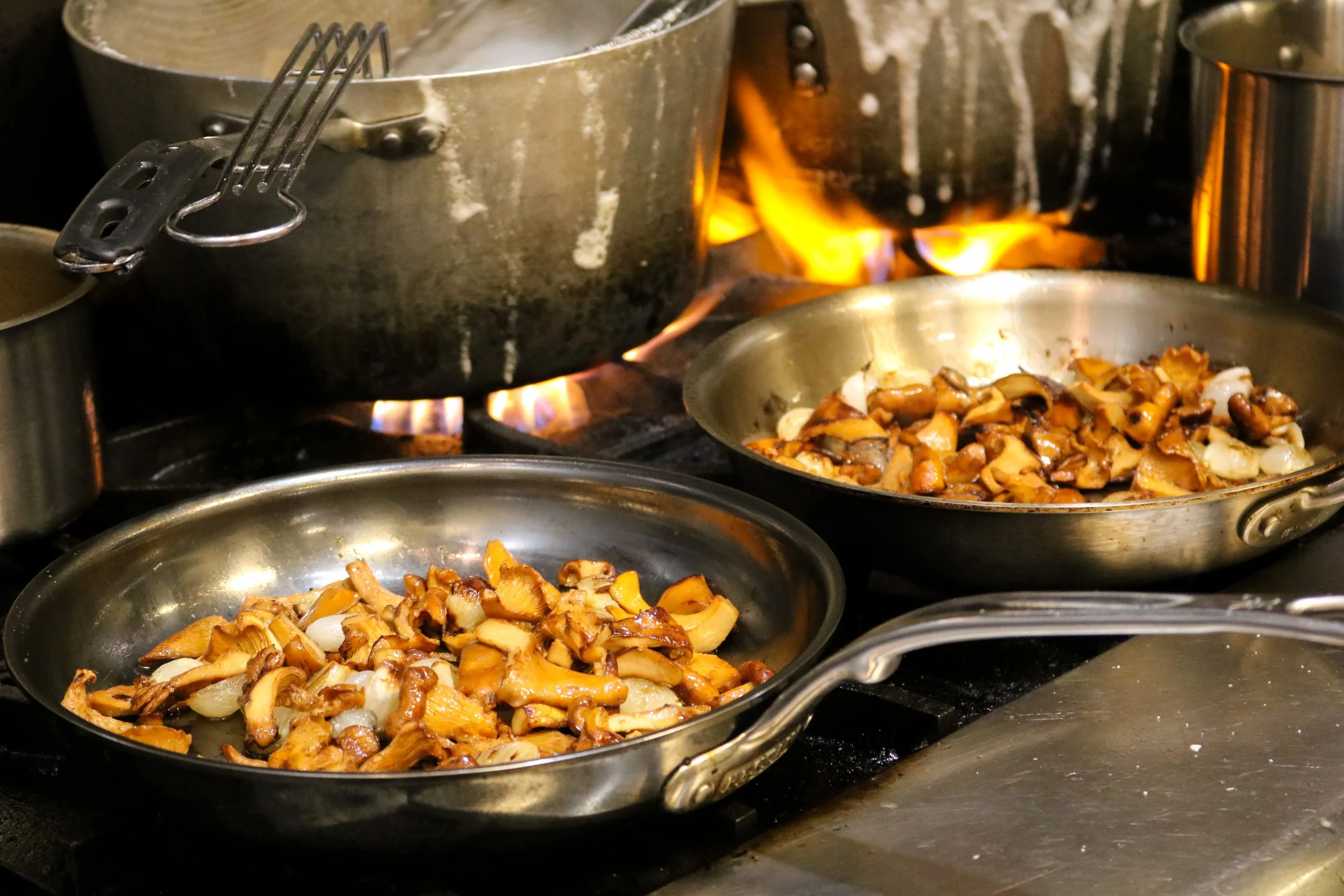 Several stainless steel pans filled with sautéed mushrooms and onions are on a stovetop, with pots and a stove flame in the background.