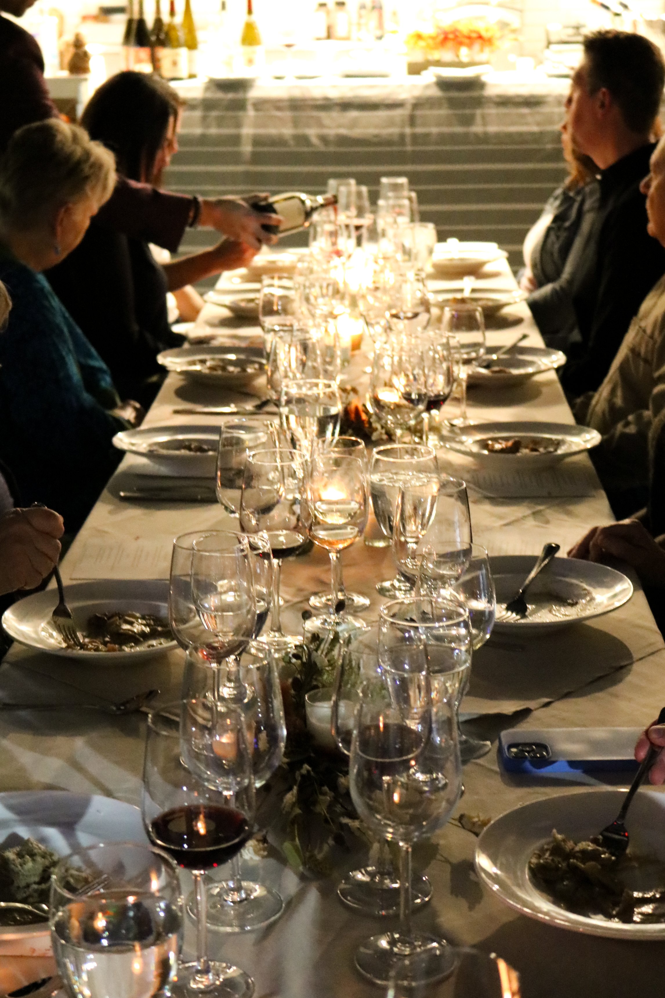 A dinner table set with multiple glasses of water and wine, plates, and utensils, in a dimly lit restaurant with a group of people gathered around.
