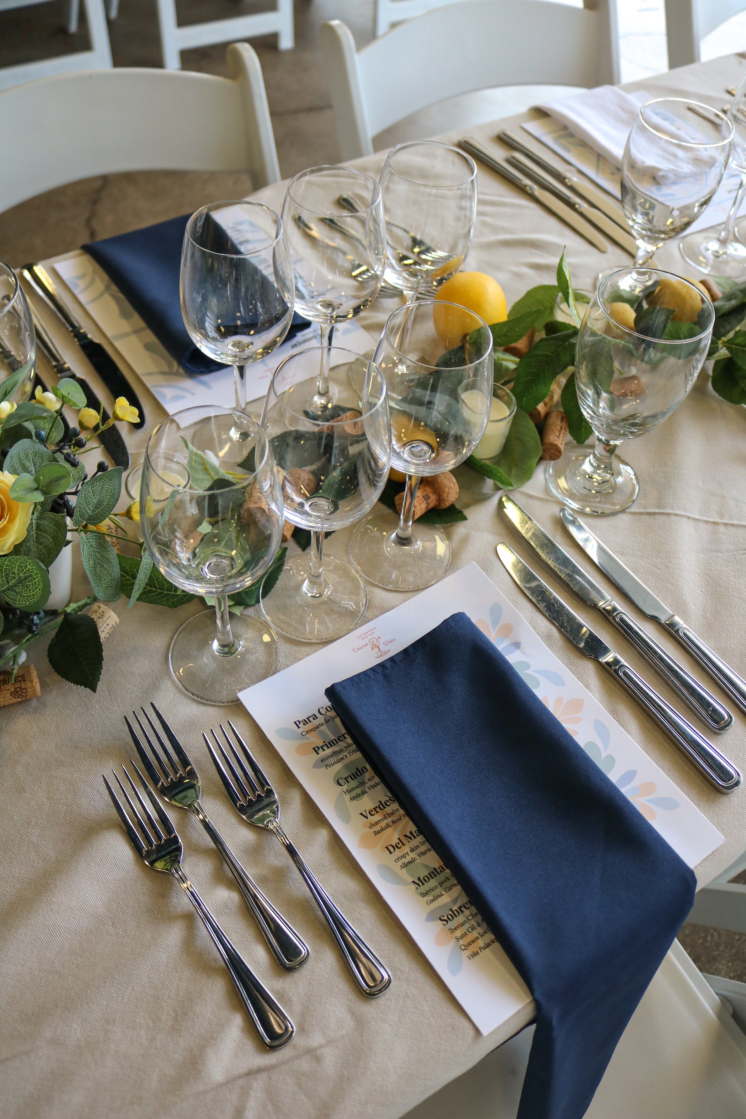 A formal dining table set with wine glasses, silverware, a navy blue napkin, and a menu. The table is decorated with yellow flowers and lemon-like fruits, with some greenery and cork accents.