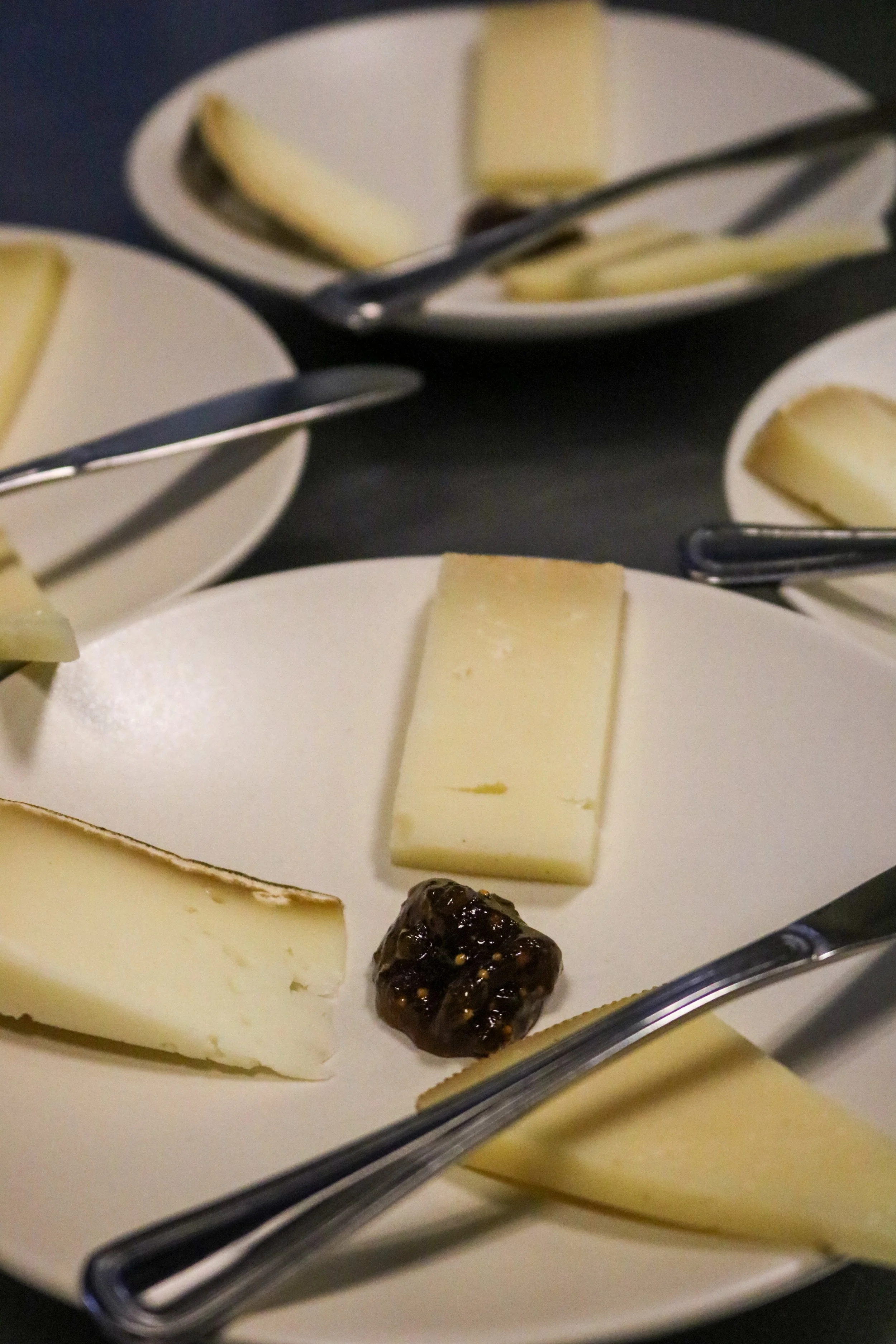 Close-up of a white cheese platter with slices of cheese, a crackled dark object, and a condiment or jam, with metal tongs resting on top.