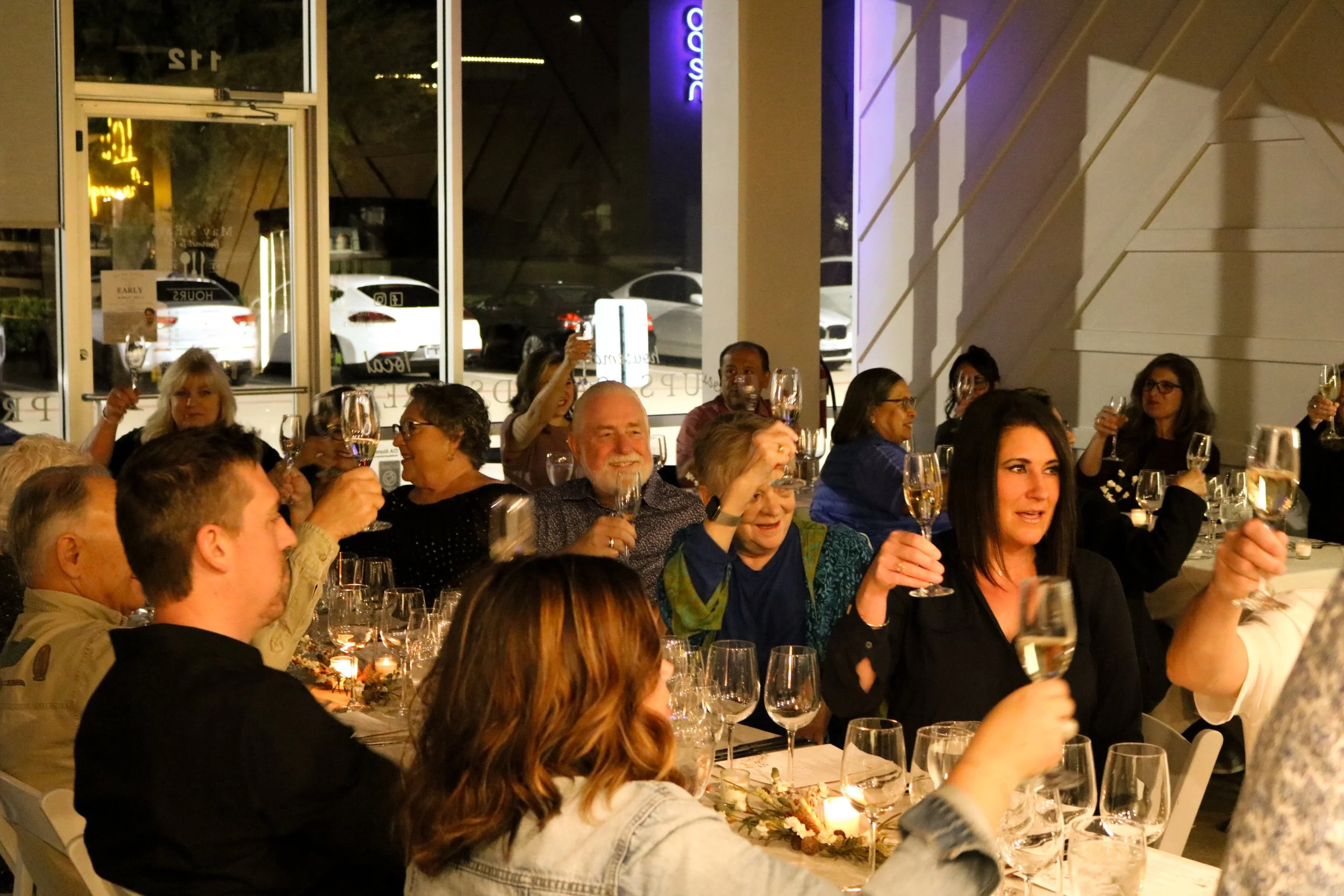 People sitting at a long dining table raising glasses in a toast during a dinner event in a restaurant, with large windows showing parked cars outside and purple neon lighting inside.