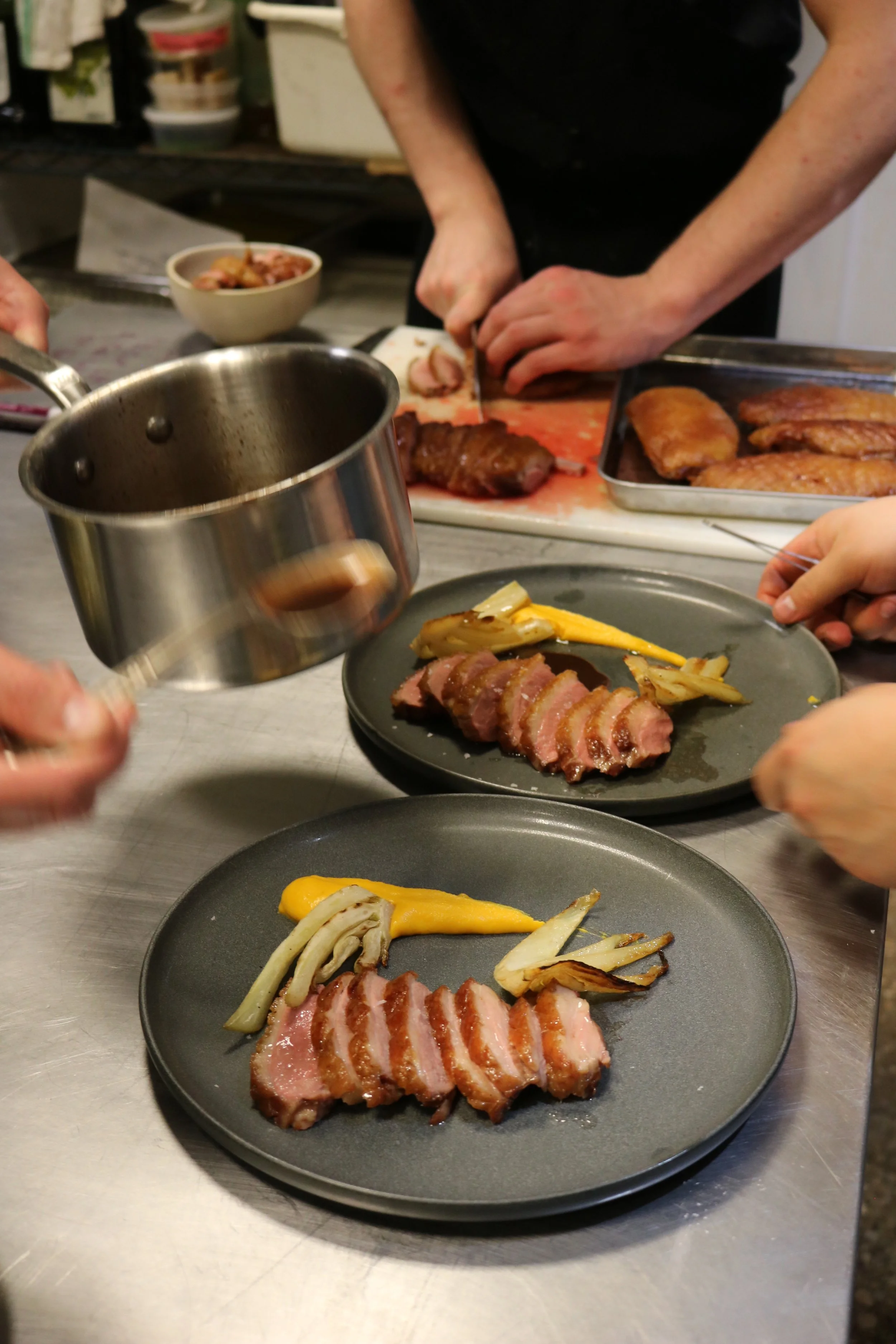 Chef slicing cooked meat on a cutting board, with plates of cooked meat and vegetables in the foreground, and other ingredients and kitchen items in the background.