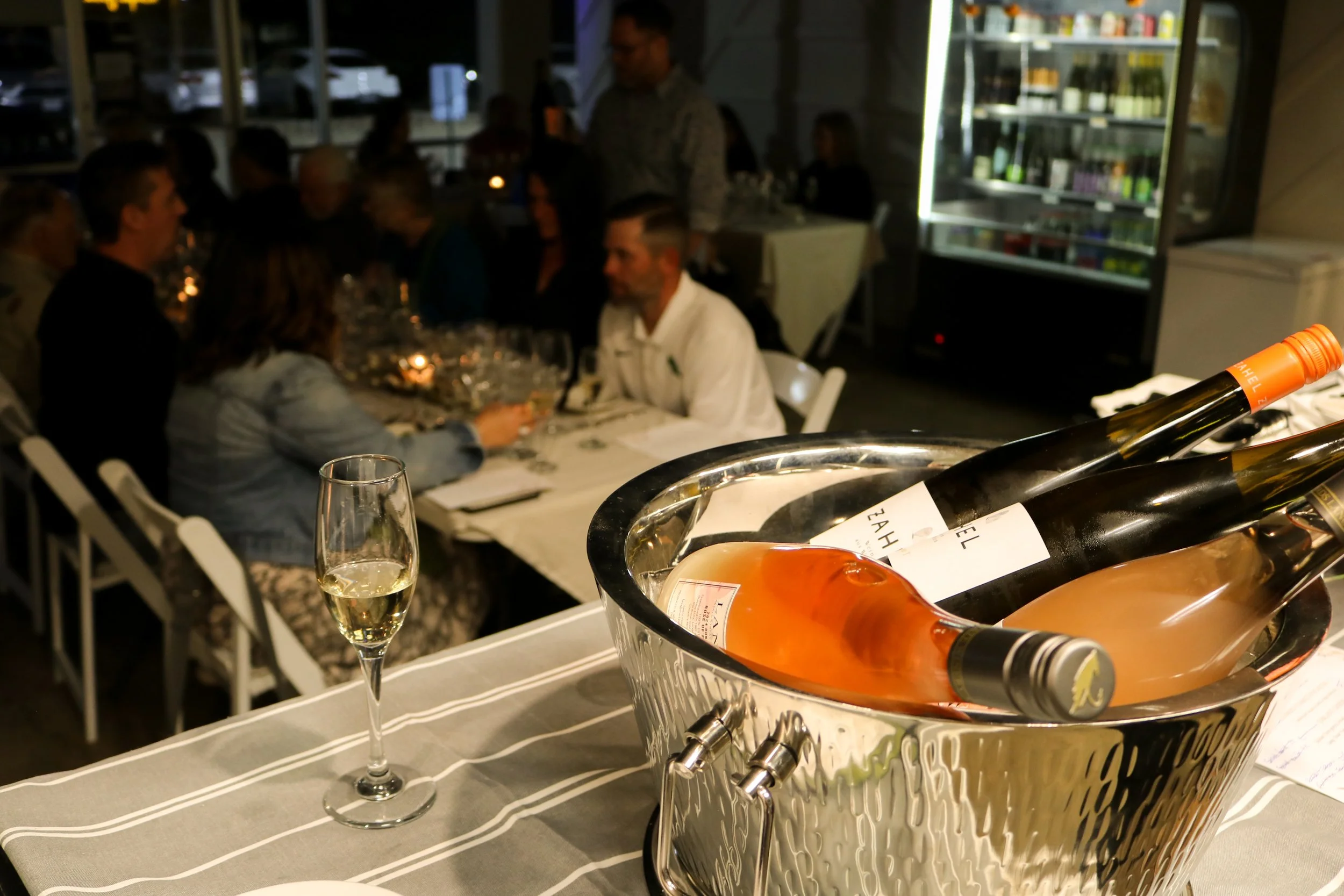A group of people dining together at a restaurant with a focus on a bucket of rosé wine bottles in the foreground. The scene is indoors, and there is a glass of white wine on the table. The setting appears to be a social dinner gathering with multipl