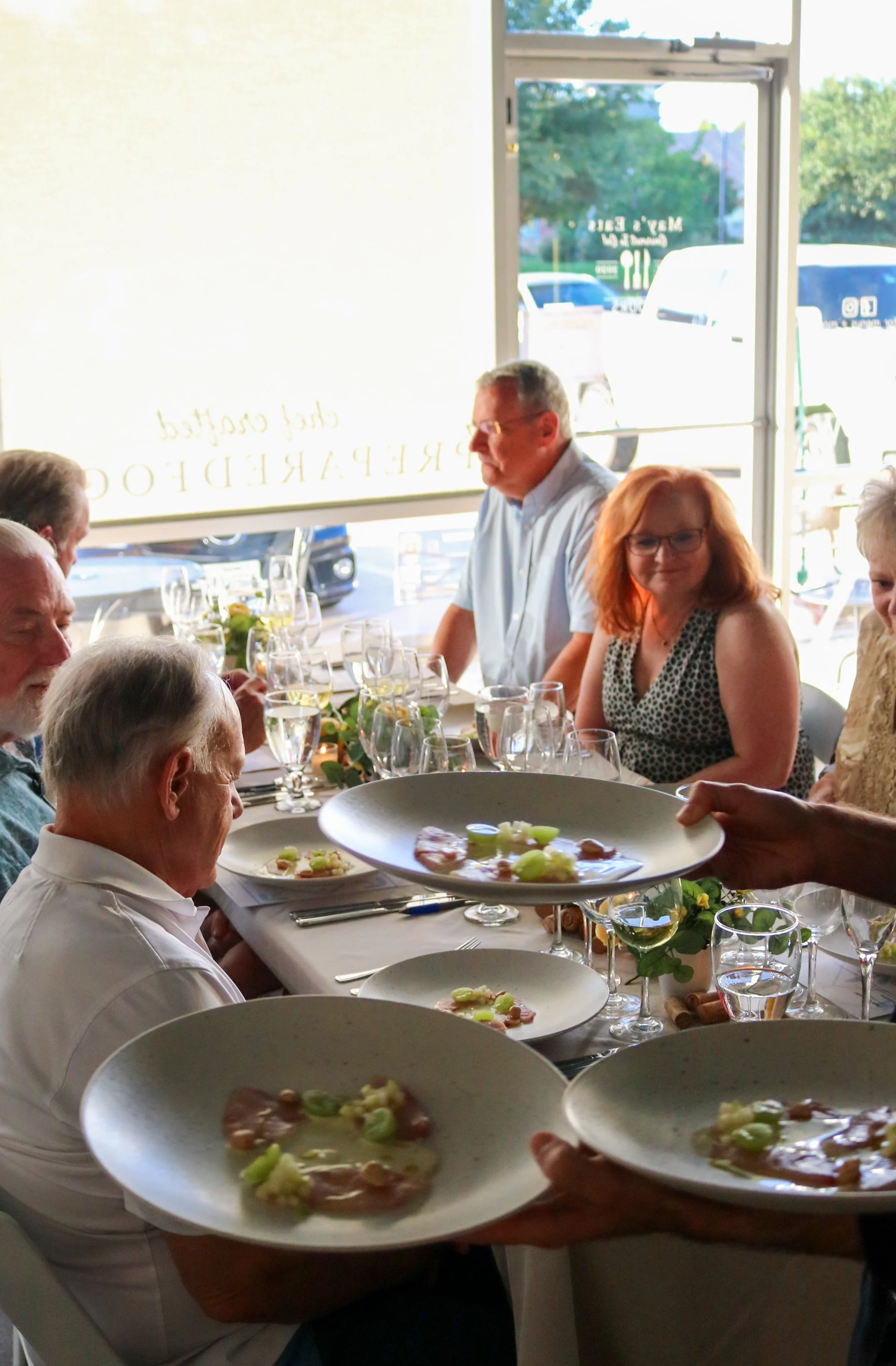 People dining inside a restaurant, being served plates of food, with a view of vehicles and trees outside through the glass door.