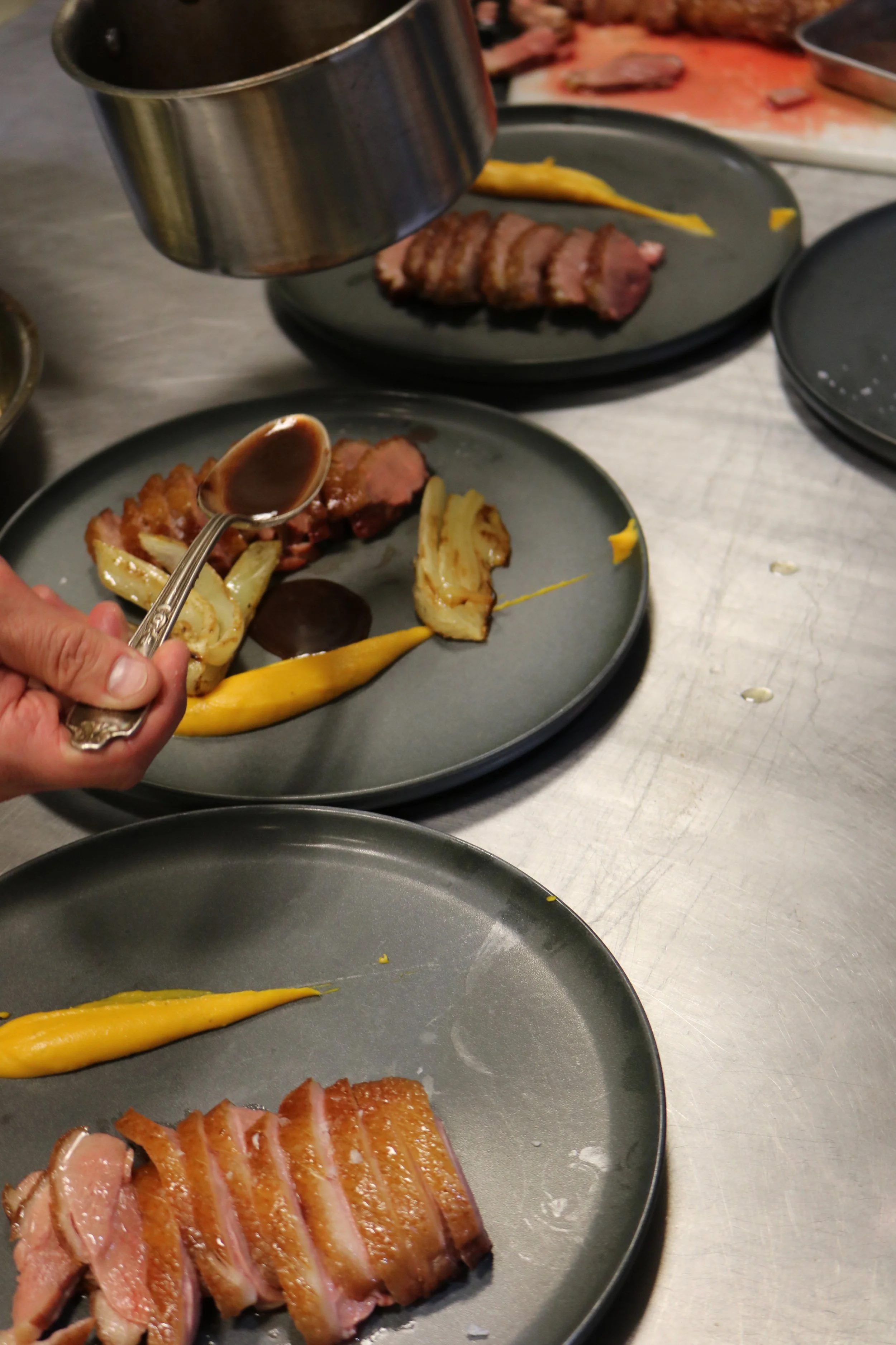 Sliced medium-rare steak with yellow sauce, topped with sauce from a spoon, on a gray plate with additional food items and plates in the background.
