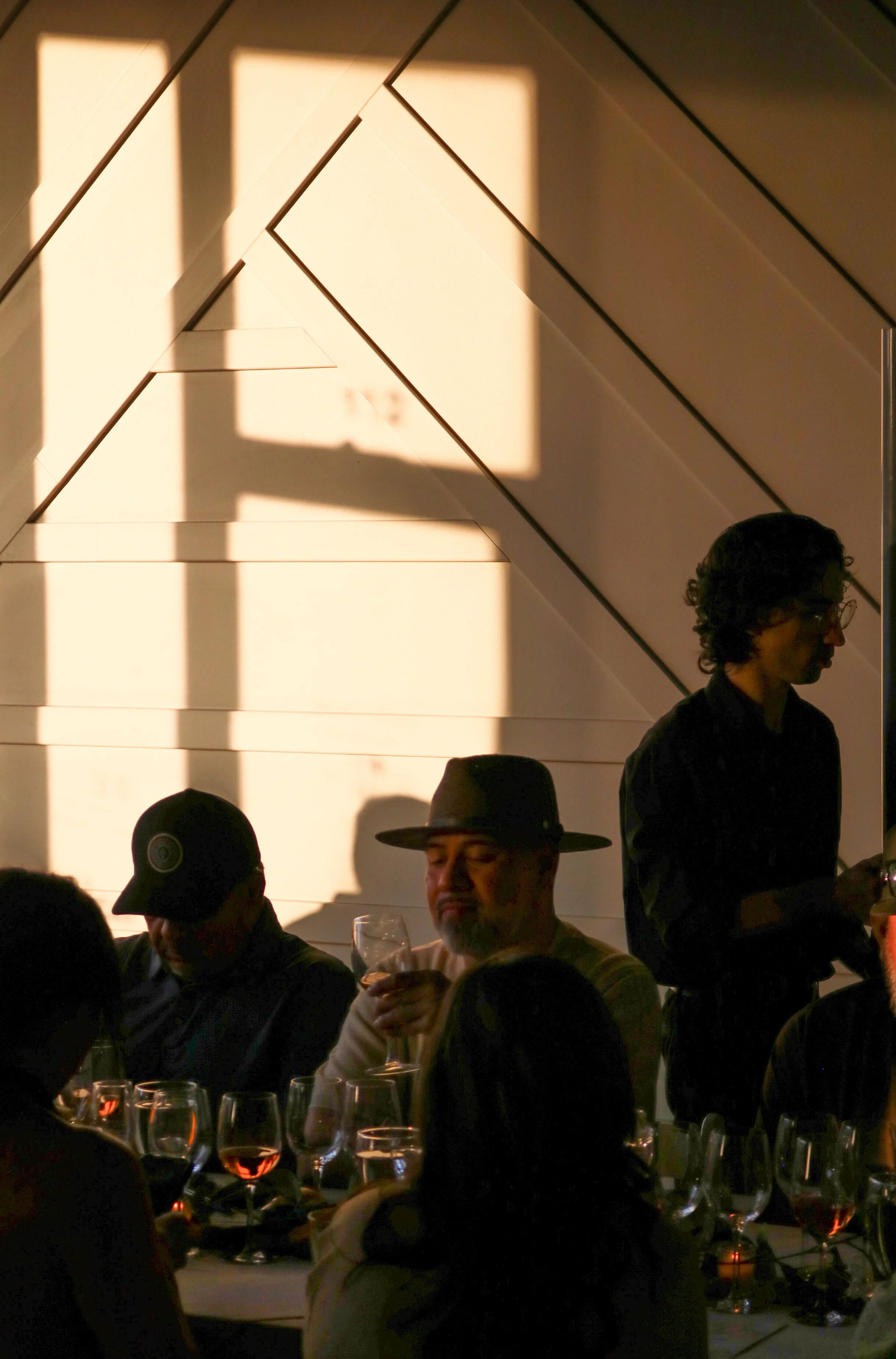 People gathered around a table with multiple glasses of red wine, with a woman standing on the right side, and a large square window casting shadow patterns on the wall behind them.