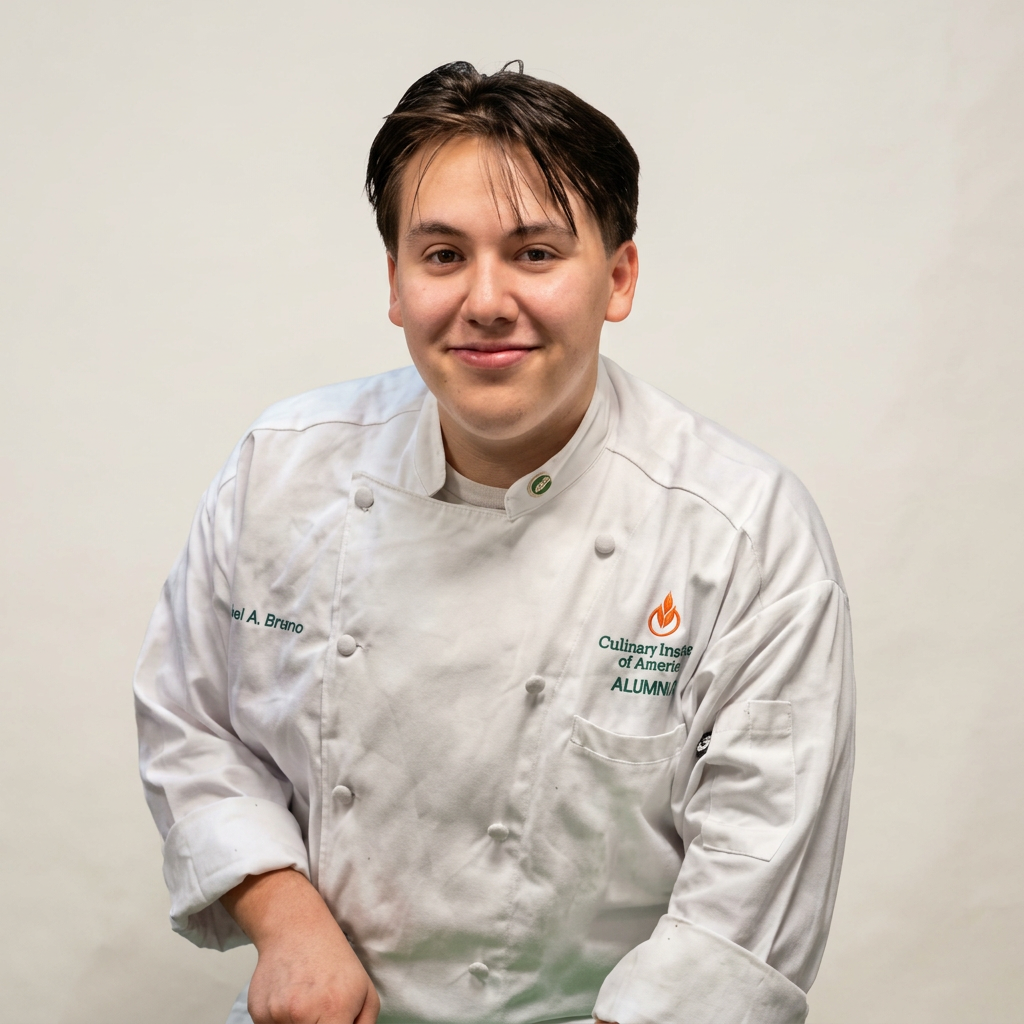 Young male chef in white uniform with 'Culinary Institute of America' logo, smiling, against plain backdrop.