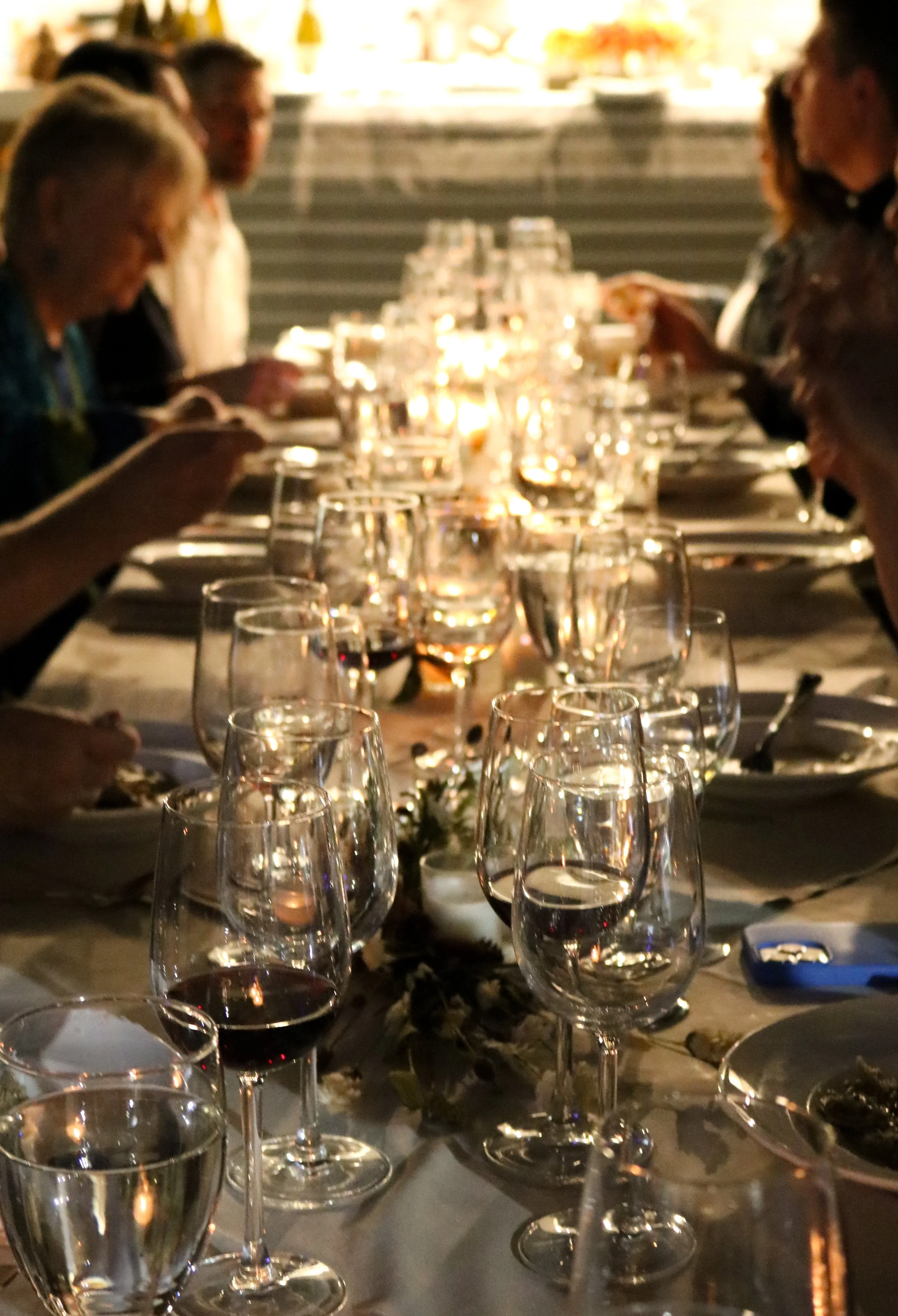 A dinner table set for a formal occasion with multiple wine glasses, some filled with red wine, and a centerpiece. People are seated around the table, engaged in conversation, with a warm, softly lit background.
