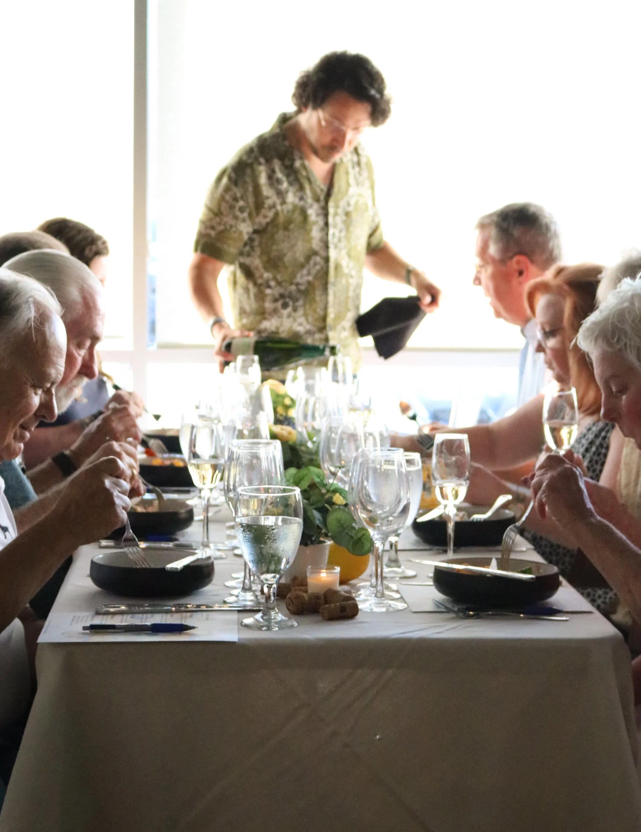 People sitting at a long dining table set for a meal, with wine glasses, candles, and a centerpiece. A man is pouring a bottle of champagne or sparkling wine, and others are eating or drinking. The setting appears to be in a bright room with large wi