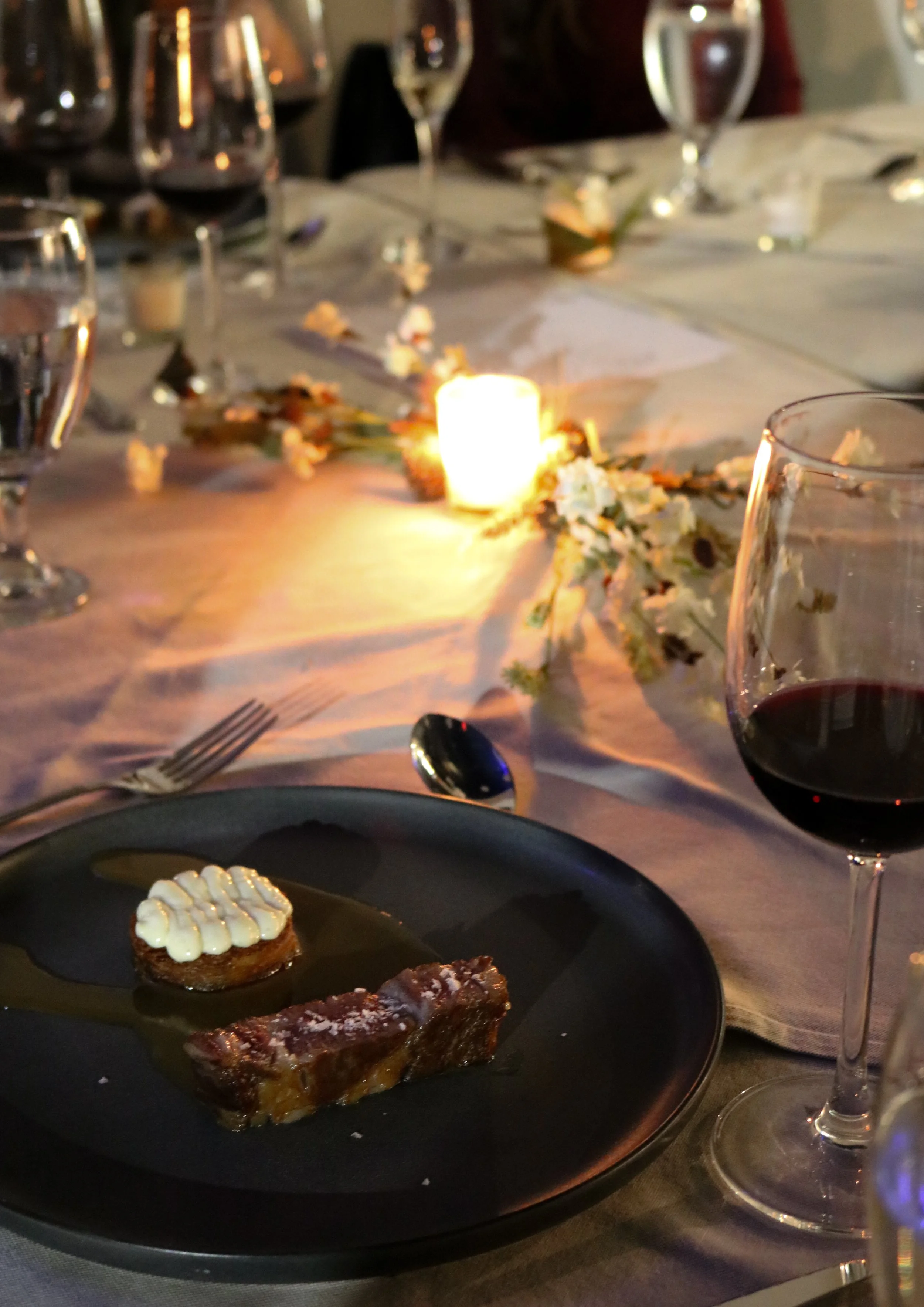 A dining table set for a meal at night, with a small lit candle, wine glasses filled with red wine, and a plate with a slice of dessert with white frosting, along with other dishes and glasses in the background.